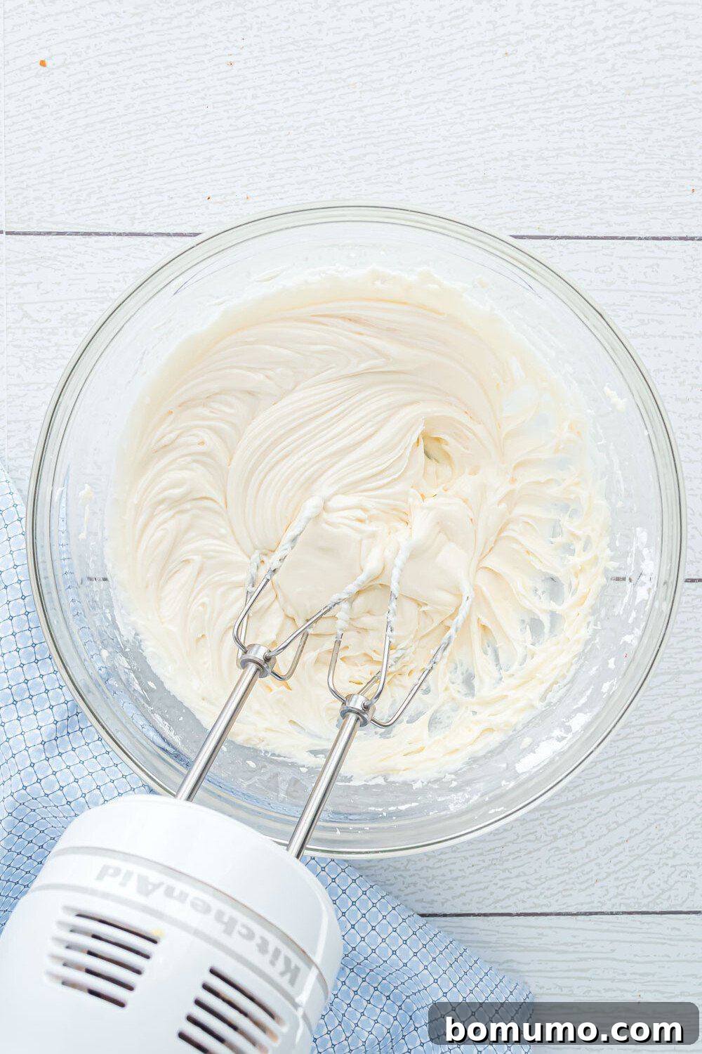 Quick Cinnamon Crescent Swirls 11 A hand mixer with beaters is resting in a glass bowl of creamy, whipped icing mixture on a light wooden surface with a blue cloth nearby.