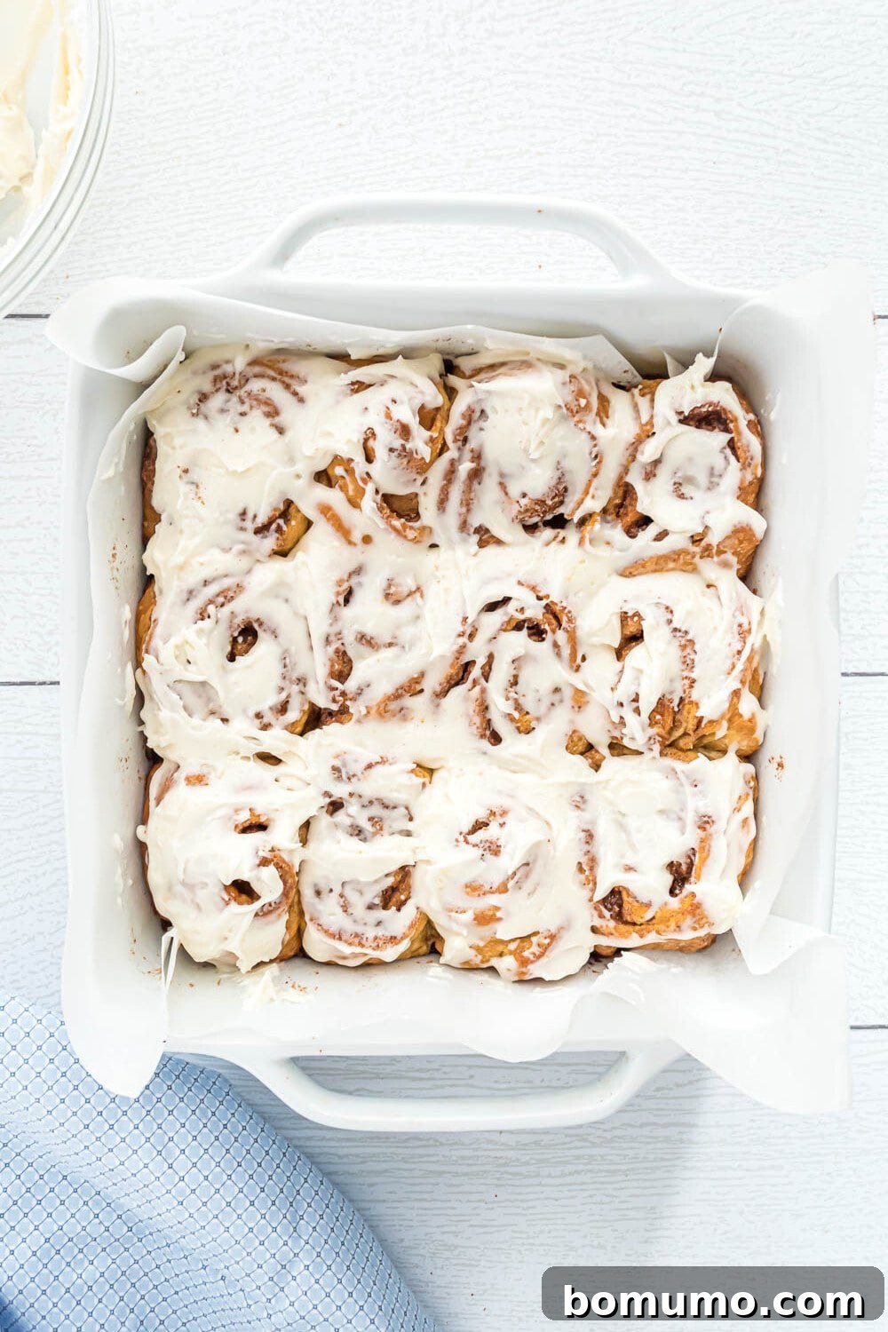 Quick Cinnamon Crescent Swirls 13 A white baking dish filled with twelve baked cinnamon rolls generously topped with luscious white icing, placed on a white surface next to a blue cloth, ready to be served.