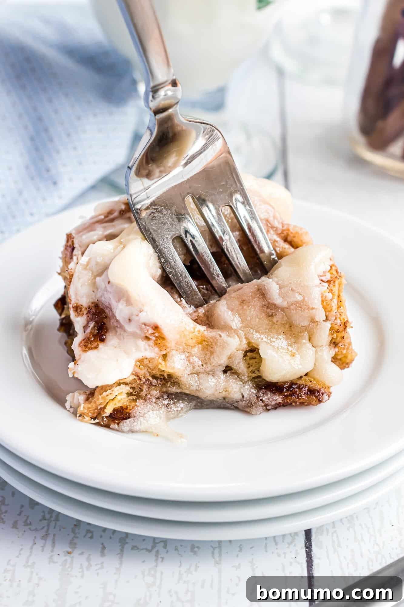 Quick Cinnamon Crescent Swirls 3 A fork cutting into a perfectly frosted crescent roll cinnamon roll, resting on a stack of white plates, showcasing its soft interior.