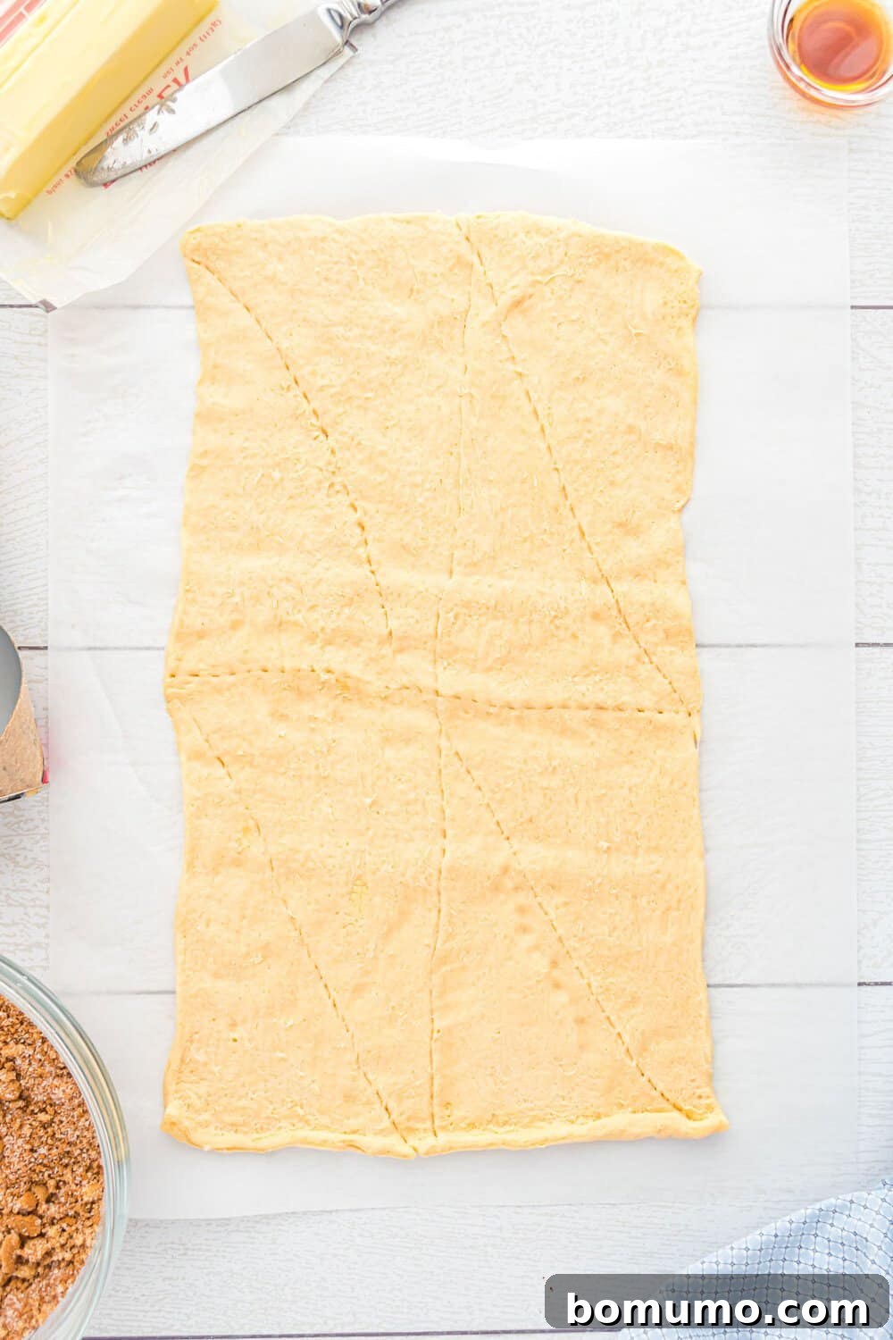 Quick Cinnamon Crescent Swirls 5 Rectangle of raw crescent dough on parchment paper, scored into triangles. Butter, a bowl of brown sugar mixture, and a knife are nearby on a light-colored surface.