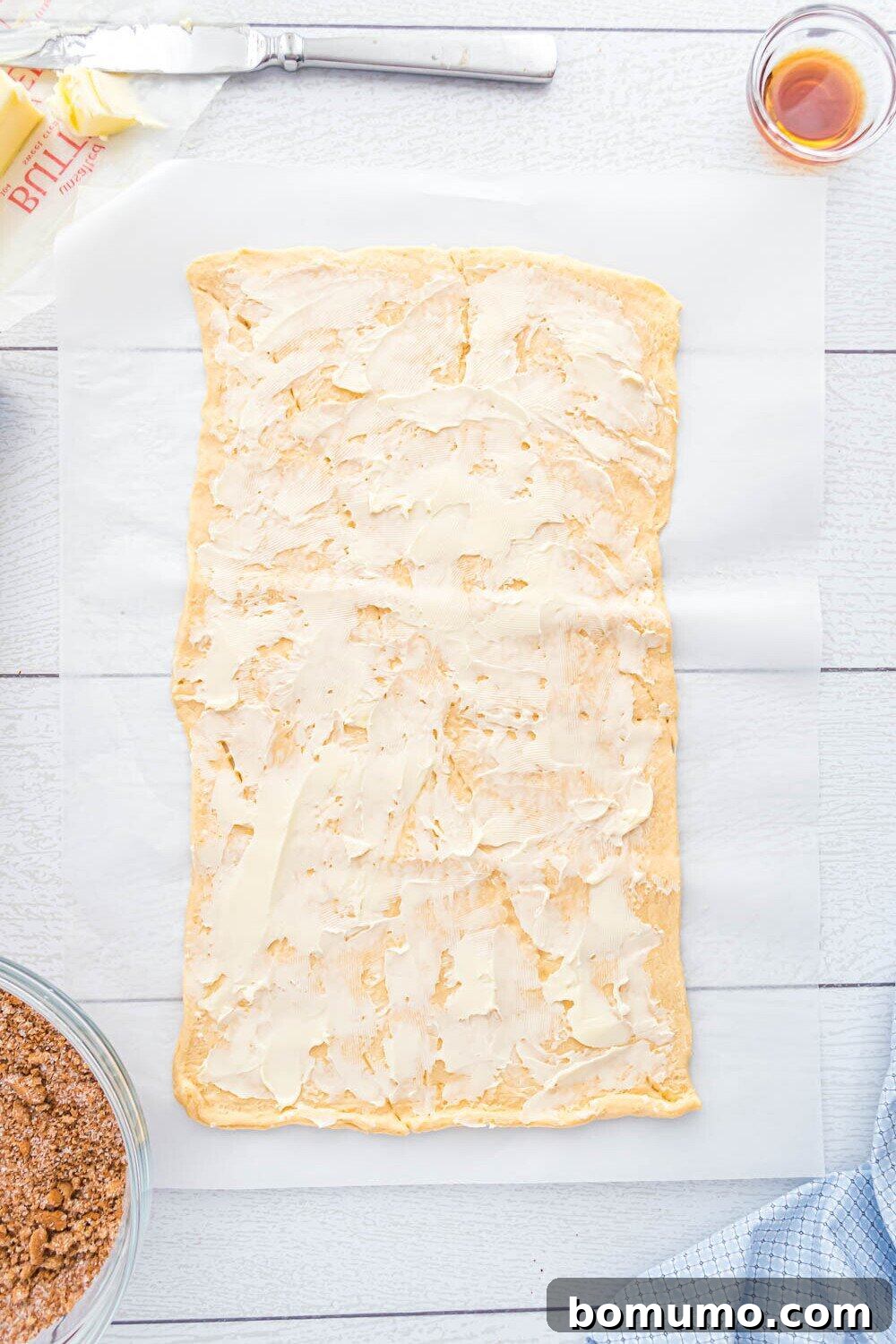 Quick Cinnamon Crescent Swirls 6 Rectangular sheet of crescent dough spread with a layer of softened butter on parchment paper, surrounded by a bowl of crumb mixture, butter, a knife, and a small cup of liquid.