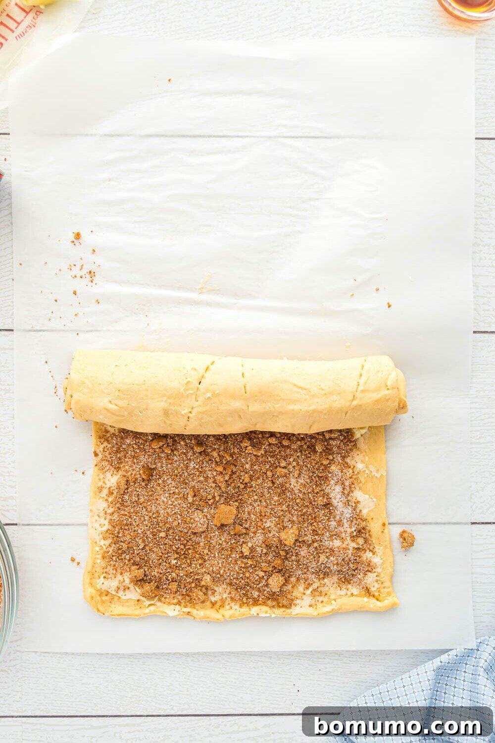 Quick Cinnamon Crescent Swirls 8 A rolled sheet of dough sprinkled with cinnamon sugar is being tightly rolled up on parchment paper on a light wooden surface.