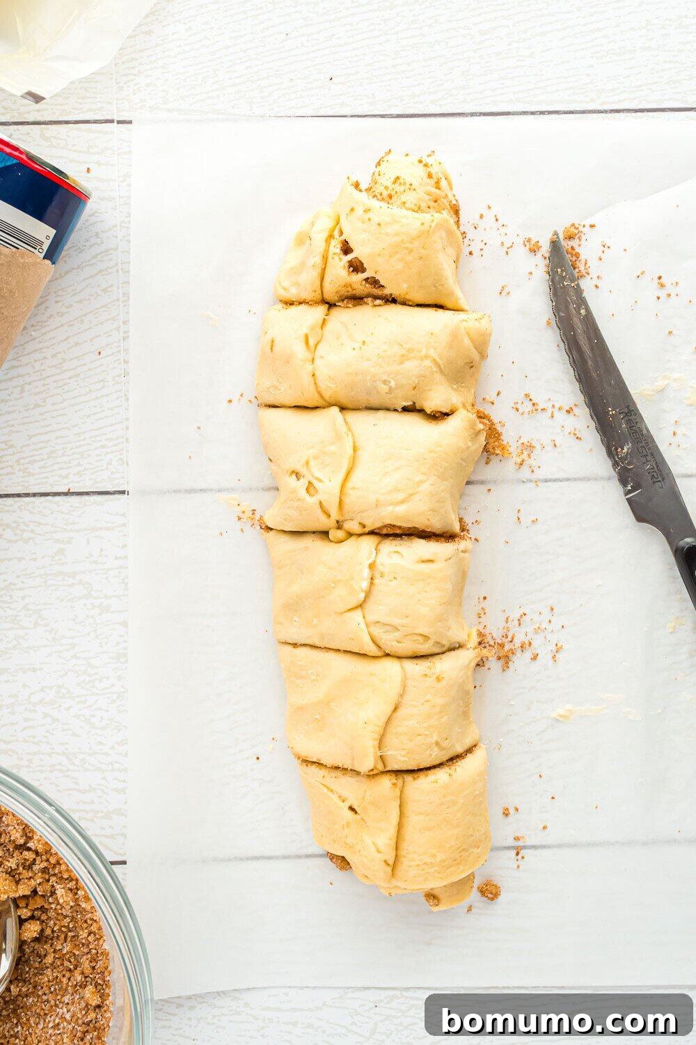 Quick Cinnamon Crescent Swirls 9 A dough log sliced into pieces on parchment paper, with a serrated knife and crumbs nearby, showcasing the distinct cinnamon swirls.
