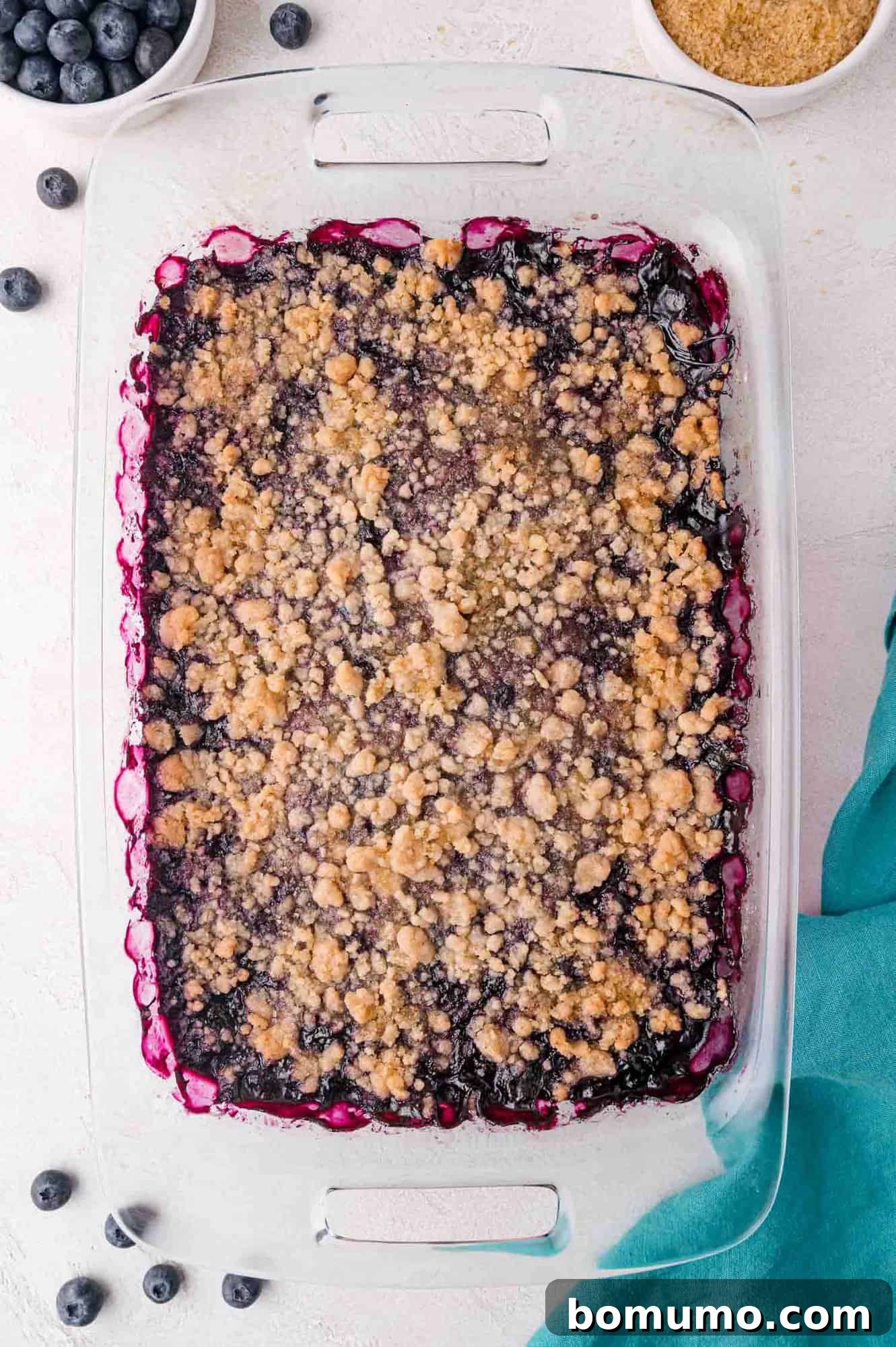 A glass baking dish filled with blueberry crumble, topped with a golden-brown streusel, sits on a light surface near a blue cloth and small bowls of blueberries and brown sugar. This image shows the perfectly baked crumble, with its inviting golden topping and rich berry filling.