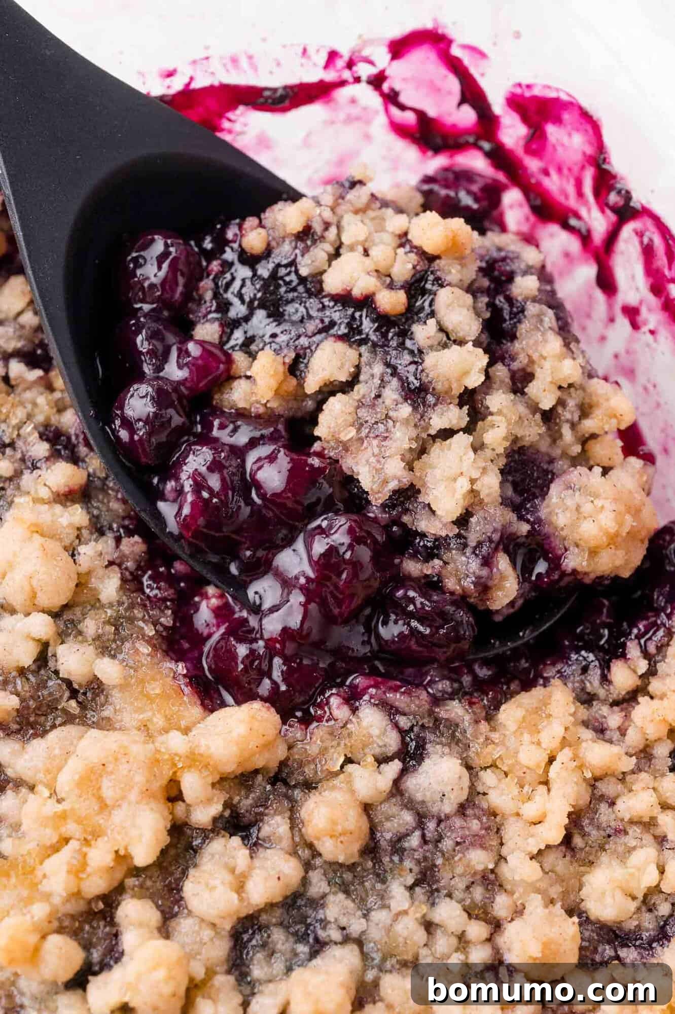 A close-up of a black spoon scooping blueberry crumble with a golden, crumbly topping and dark purple blueberry filling. This image emphasizes the tempting textures of the dessert.