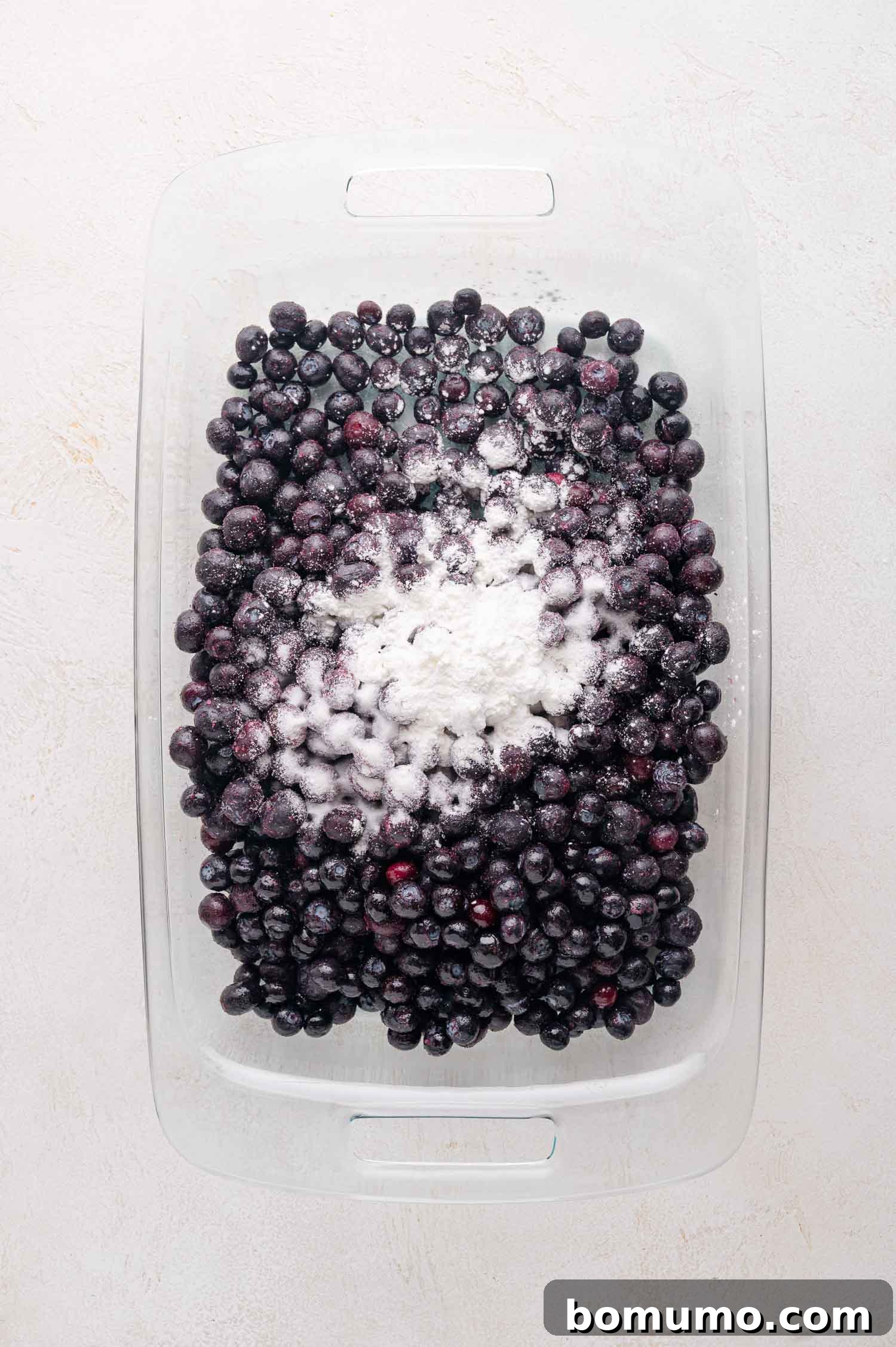 A glass baking dish filled with blueberries topped with a mound of cornstarch and sugar. This photo illustrates the initial step of preparing the blueberry filling, showing the raw ingredients before mixing.