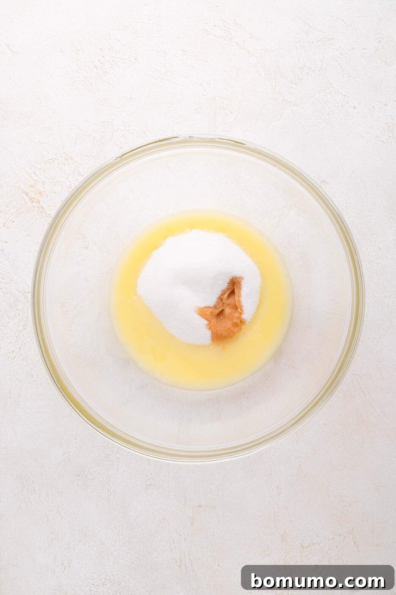 A glass mixing bowl containing melted butter, granulated sugar, and vanilla on a light-colored surface. This shows the initial wet ingredients for the crumble topping.