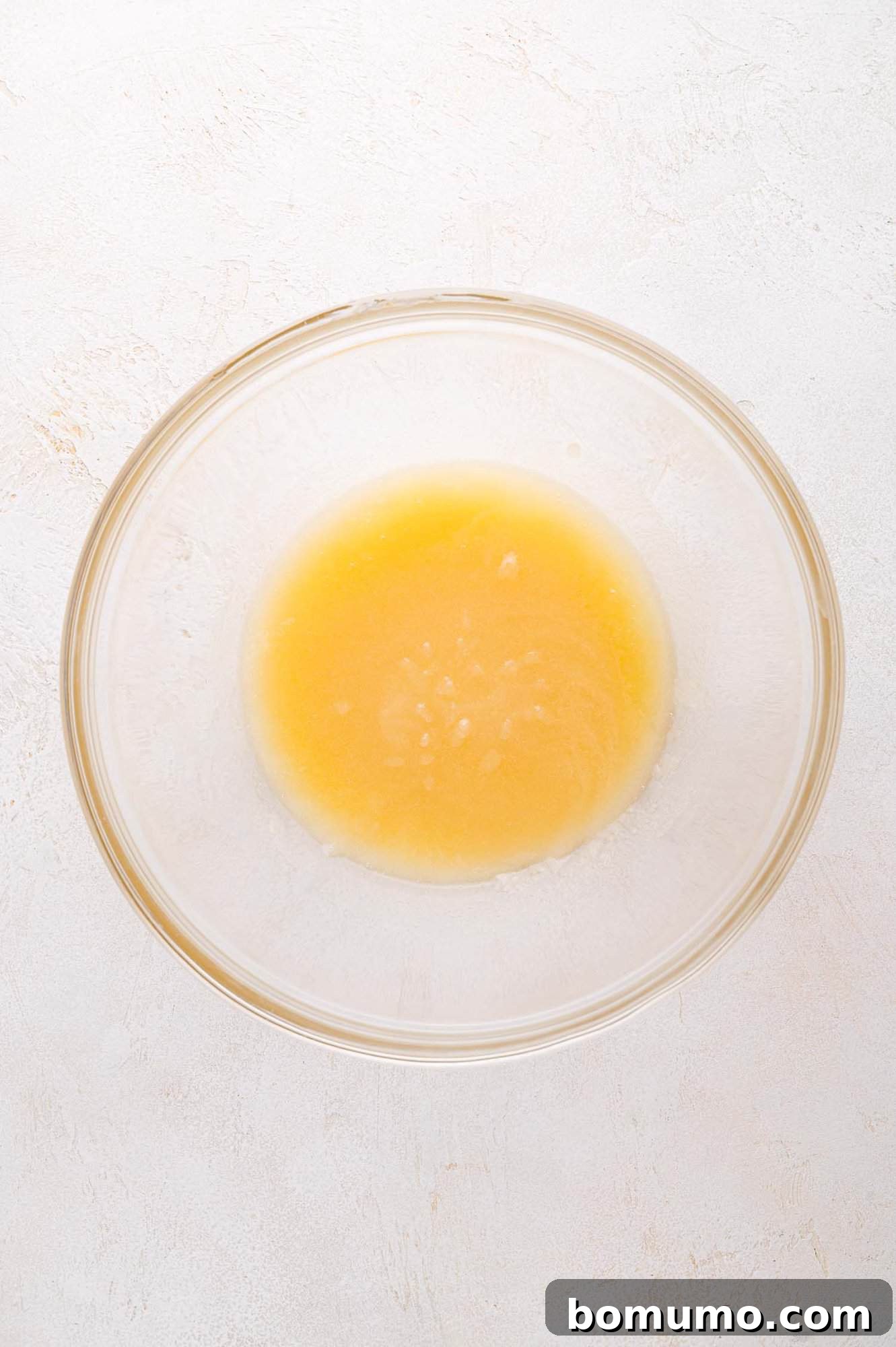 A glass bowl containing a small amount of yellow liquid mixture (butter, sugar, vanilla) sits on a light-colored surface. This highlights the blended wet ingredients, ready for the dry components.