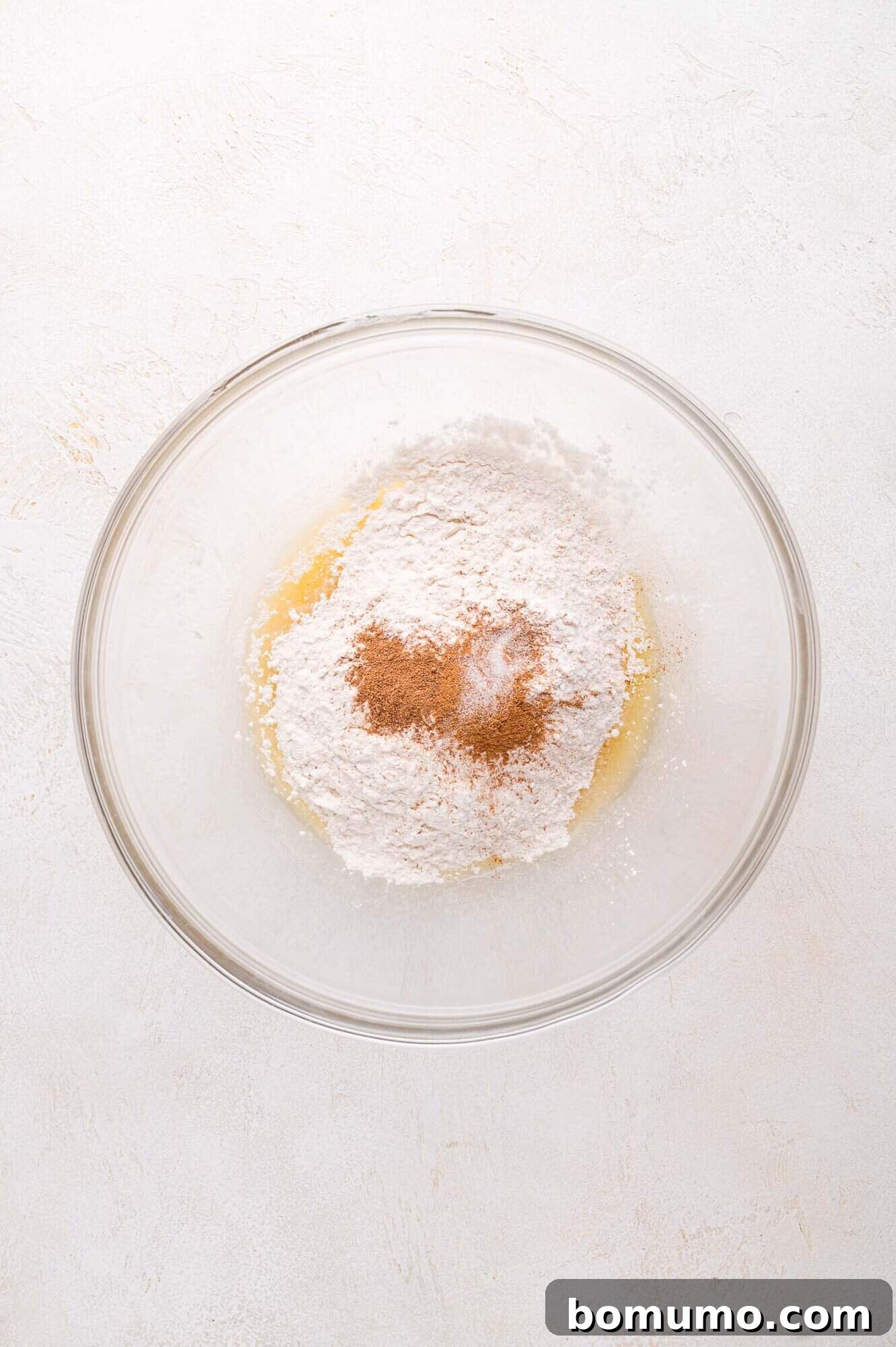 A glass bowl containing flour, cinnamon, and a wet ingredient mixture on a light-colored surface. This image demonstrates the addition of dry ingredients to the wet mixture, a key step in creating the crumble.