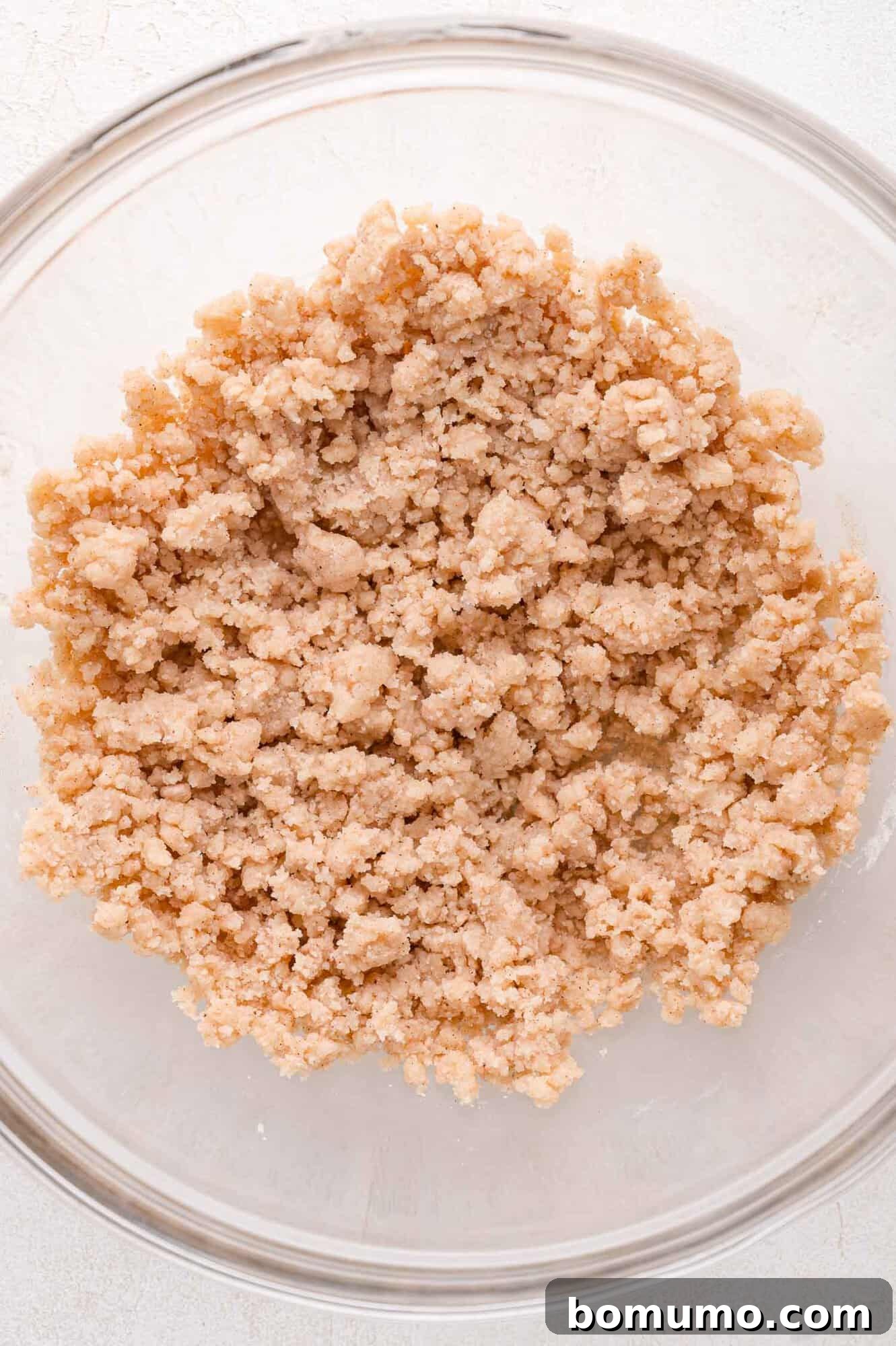 A clear glass bowl containing a crumbly, light brown mixture on a white surface. This final image in the sequence shows the perfectly mixed crumble topping, ready to be spread over the blueberries.