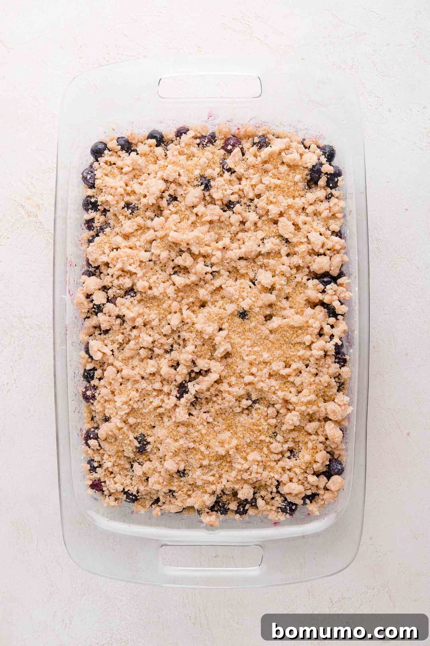 A rectangular glass baking dish filled with blueberry crumble topped with a layer of crumbly, golden-brown streusel, ready to be baked. This image captures the delicious anticipation of the dessert before it goes into the oven.