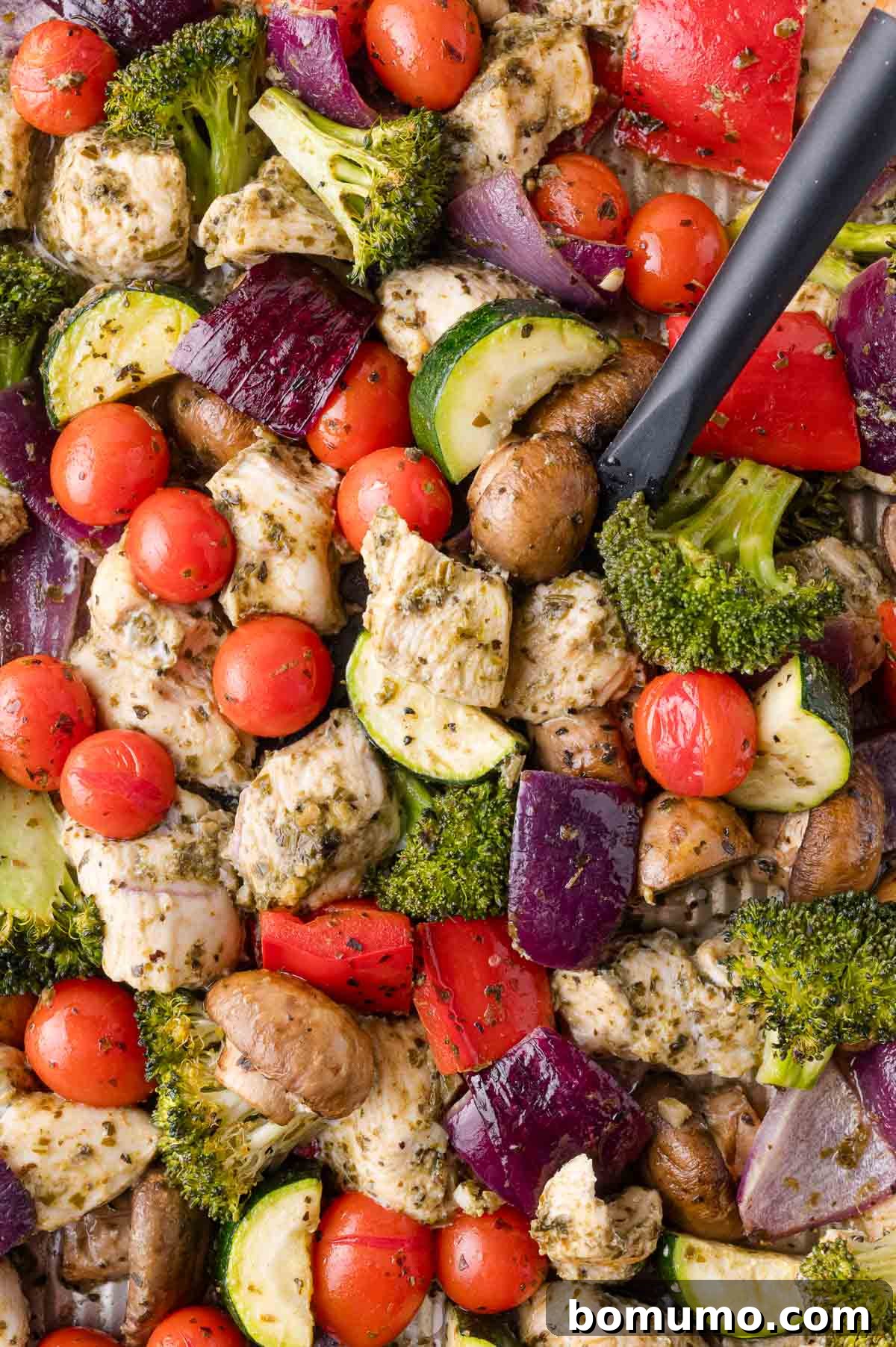 A colorful sheet pan loaded with perfectly baked pesto chicken cubes and an array of roasted vegetables like broccoli florets, red onion, bell peppers, mushrooms, zucchini slices, and cherry tomatoes, fresh out of the oven. A black spatula is resting on the right side of the pan, ready for serving.