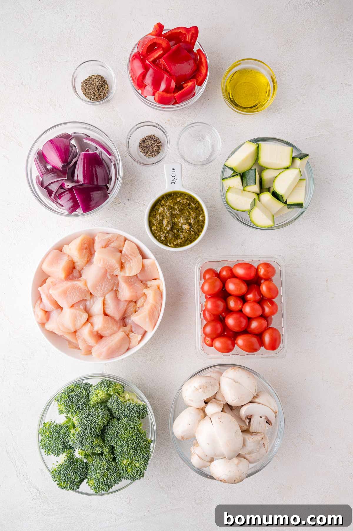 Assorted bowls showcasing the fresh ingredients: raw chicken, vibrant broccoli florets, sliced mushrooms, plump grape tomatoes, zucchini rounds, chunky red onion, red bell pepper strips, a bowl of rich green pesto, a small dish of olive oil, and pinches of salt and pepper, all neatly arranged on a pristine white surface, ready for preparation.