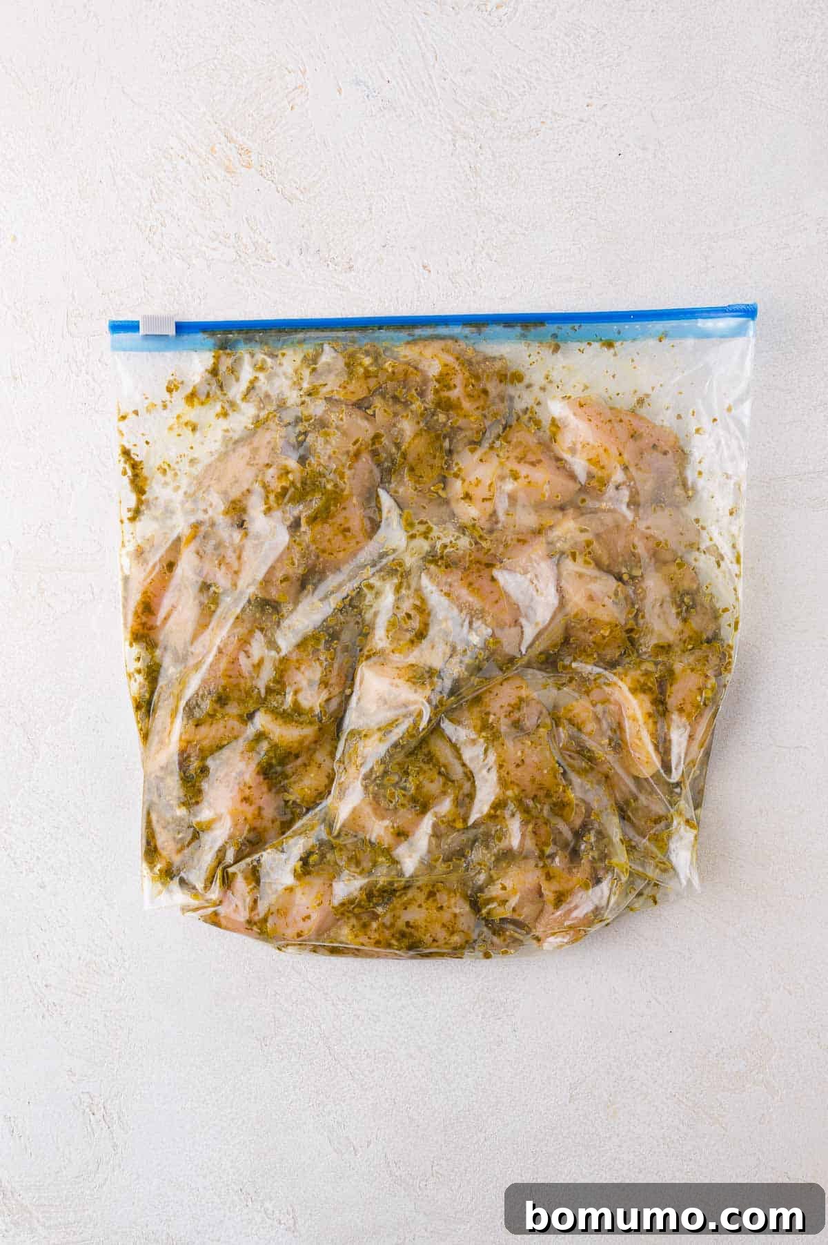 Close-up shot of raw chicken pieces marinating in a clear resealable plastic bag, coated generously with a bright green pesto sauce. The bag is resting on a light-colored countertop, showcasing the marinade process for the Sheet Pan Pesto Chicken.