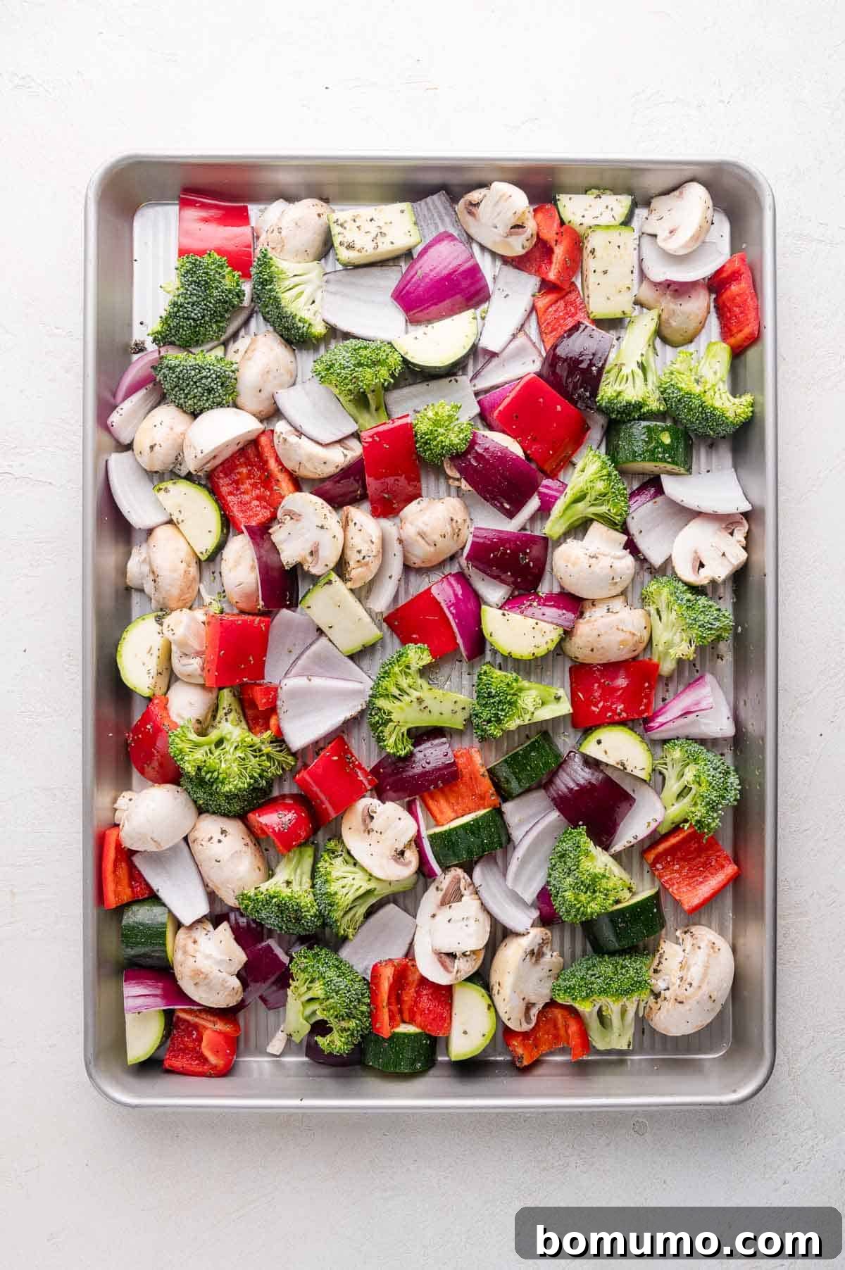 A pristine baking sheet evenly covered with chopped vegetables including earthy mushrooms, vibrant broccoli florets, chunky red onion, delicate zucchini slices, and crisp red bell pepper pieces, all lightly seasoned with herbs and freshly ground black pepper, ready for the oven to begin roasting.