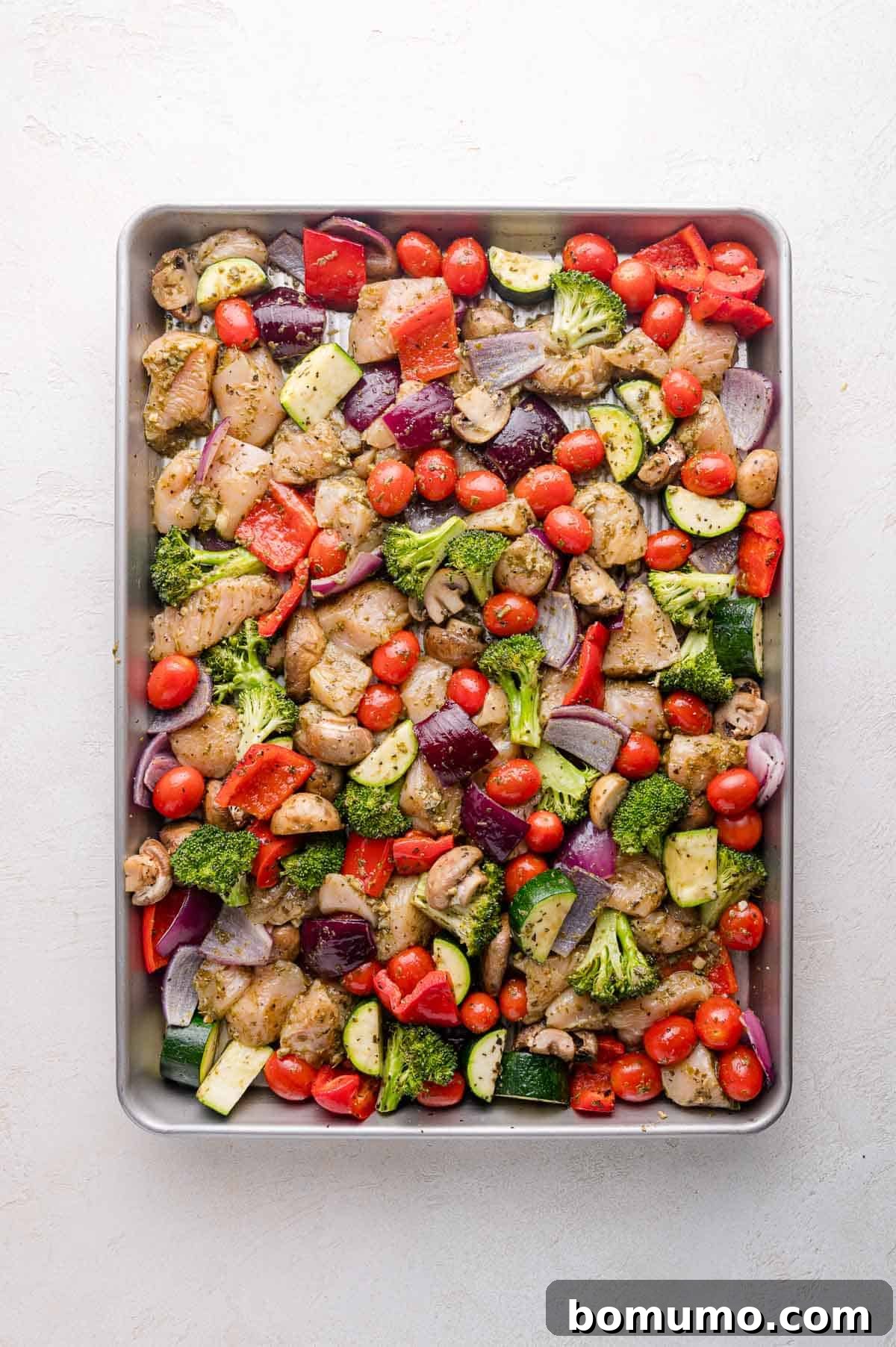An overhead view of a large baking tray brimming with a delicious assortment of cubed chicken, vibrant broccoli florets, zucchini slices, bright red bell peppers, juicy cherry tomatoes, and chunky red onions. Everything is generously coated and seasoned, spread out in a single layer, poised for roasting to create a perfect Sheet Pan Pesto Chicken meal.