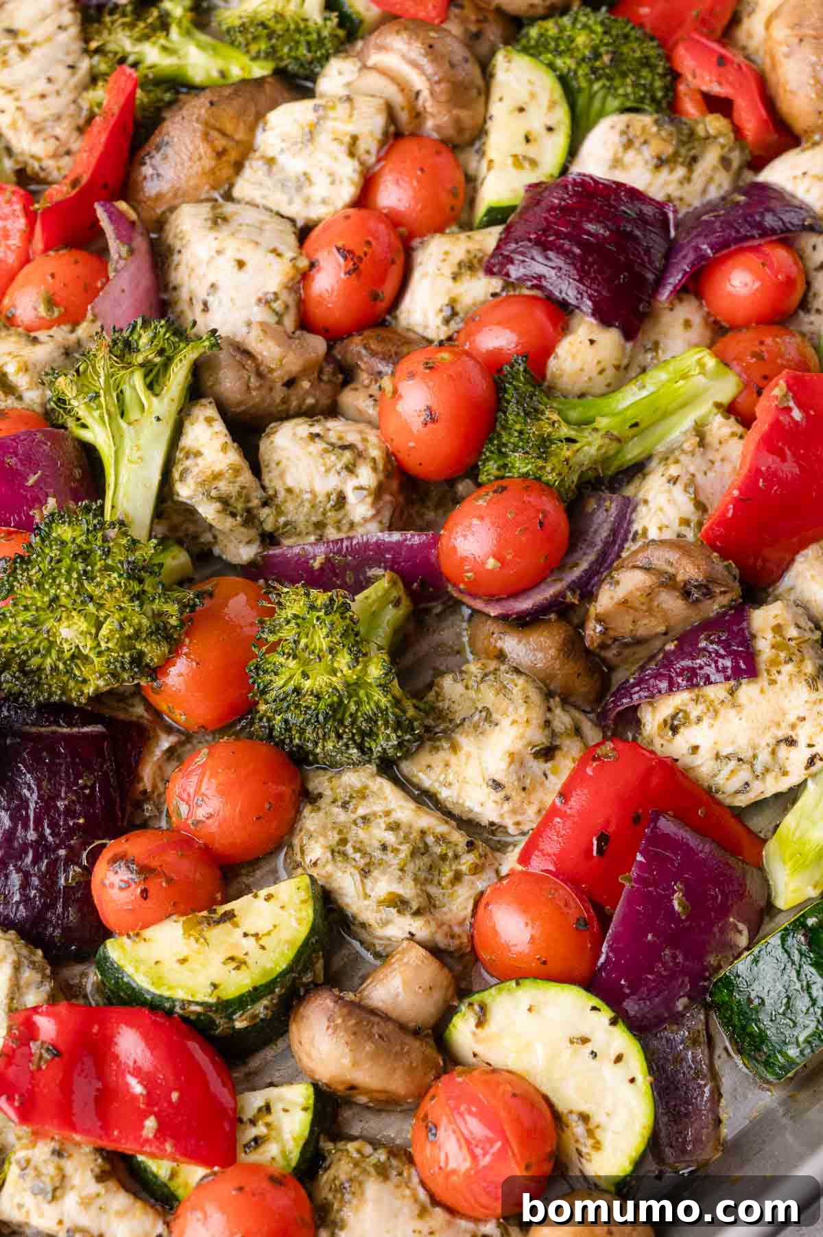 A beautifully cooked sheet pan filled with golden-brown chicken pieces, glistening cherry tomatoes, tender broccoli florets, soft zucchini, sautéed mushrooms, caramelized red onion, and crisp red bell peppers, all perfectly seasoned with aromatic herbs, making for a hearty and complete meal.