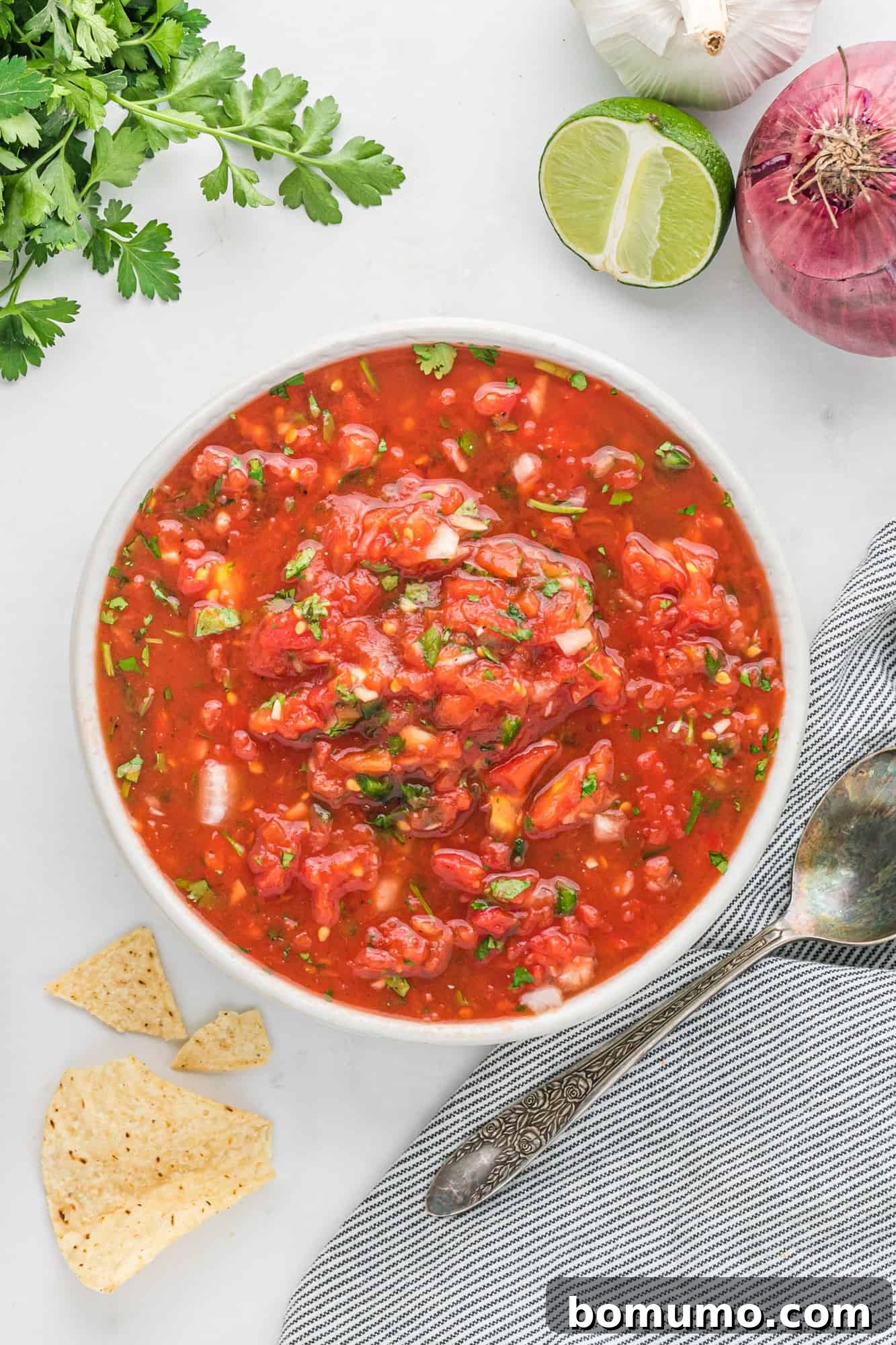 A bowl of vibrant homemade salsa surrounded by tortilla chips, fresh cilantro, and lime wedges.