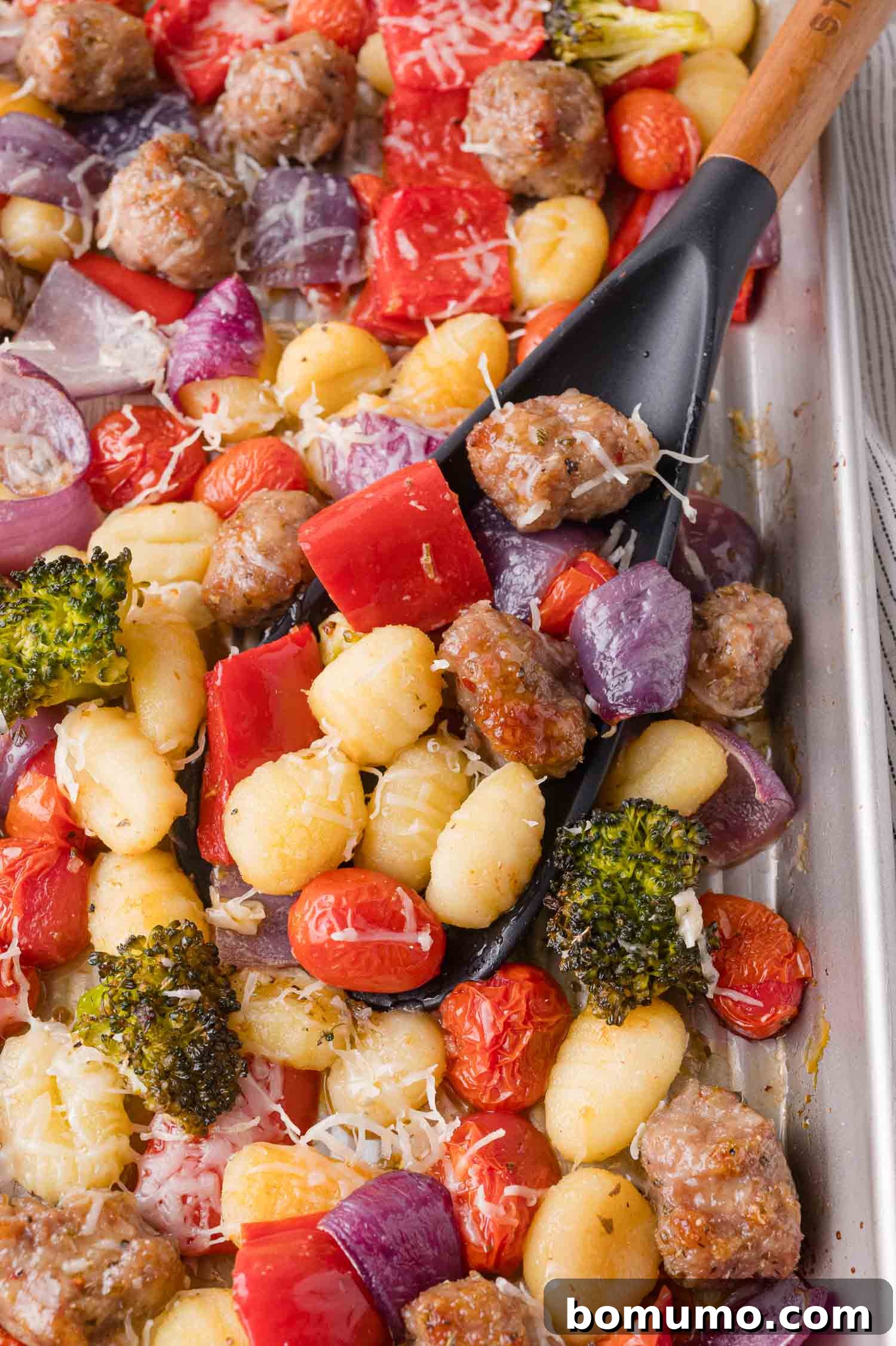A sheet pan with cooked gnocchi, Italian sausage, cherry tomatoes, red onion, red bell pepper, broccoli, and a sprinkle of shredded cheese, with a black spatula scooping a portion.