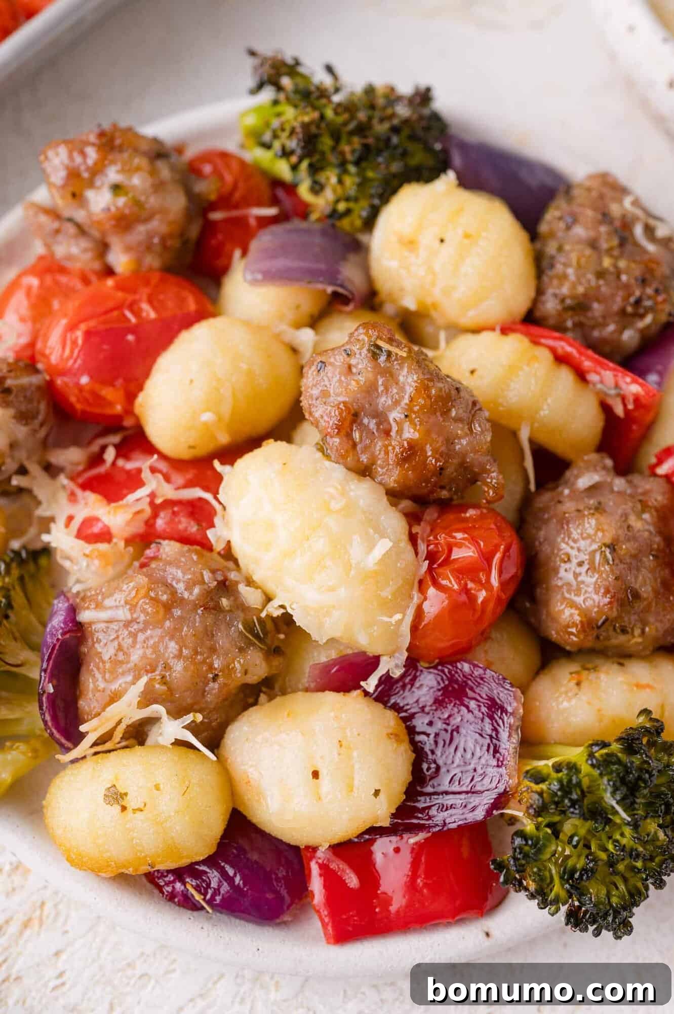 A neatly arranged plate of sheet pan gnocchi, featuring crispy gnocchi, savory Italian sausage, vibrant cherry tomatoes, caramelized red onion, tender red bell peppers, and broccoli, all finished with a light dusting of grated Parmesan cheese.