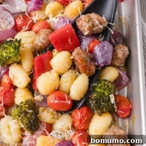A tray of baked gnocchi with sausage, cherry tomatoes, broccoli, red onions, red bell peppers, and grated cheese, with a black serving spoon.