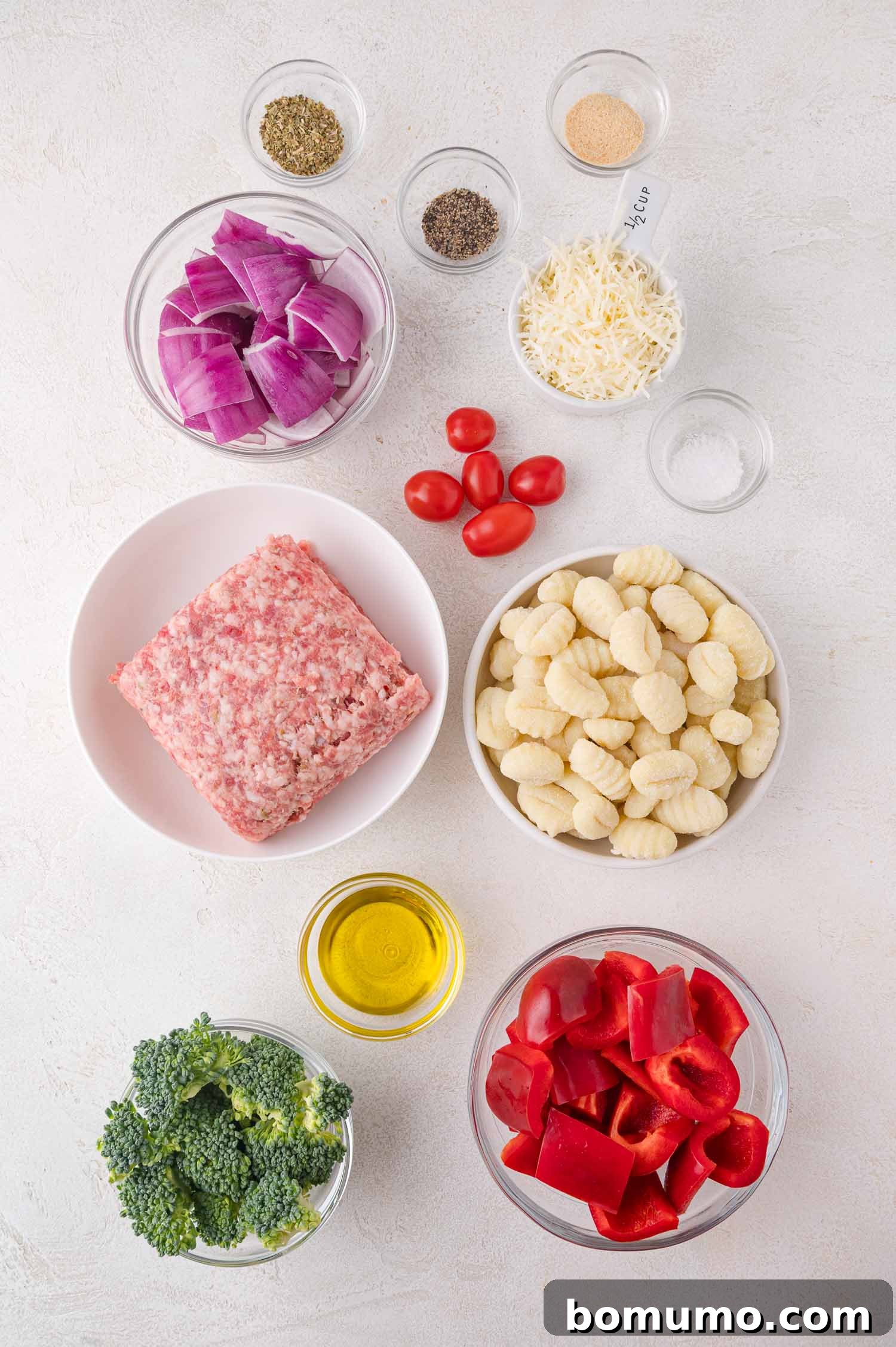 Assorted ingredients for sheet pan gnocchi on a white surface, including raw ground Italian sausage, uncooked gnocchi, broccoli florets, red bell pepper chunks, cherry tomatoes, red onion pieces, shredded Parmesan cheese, and small bowls of olive oil and spices.