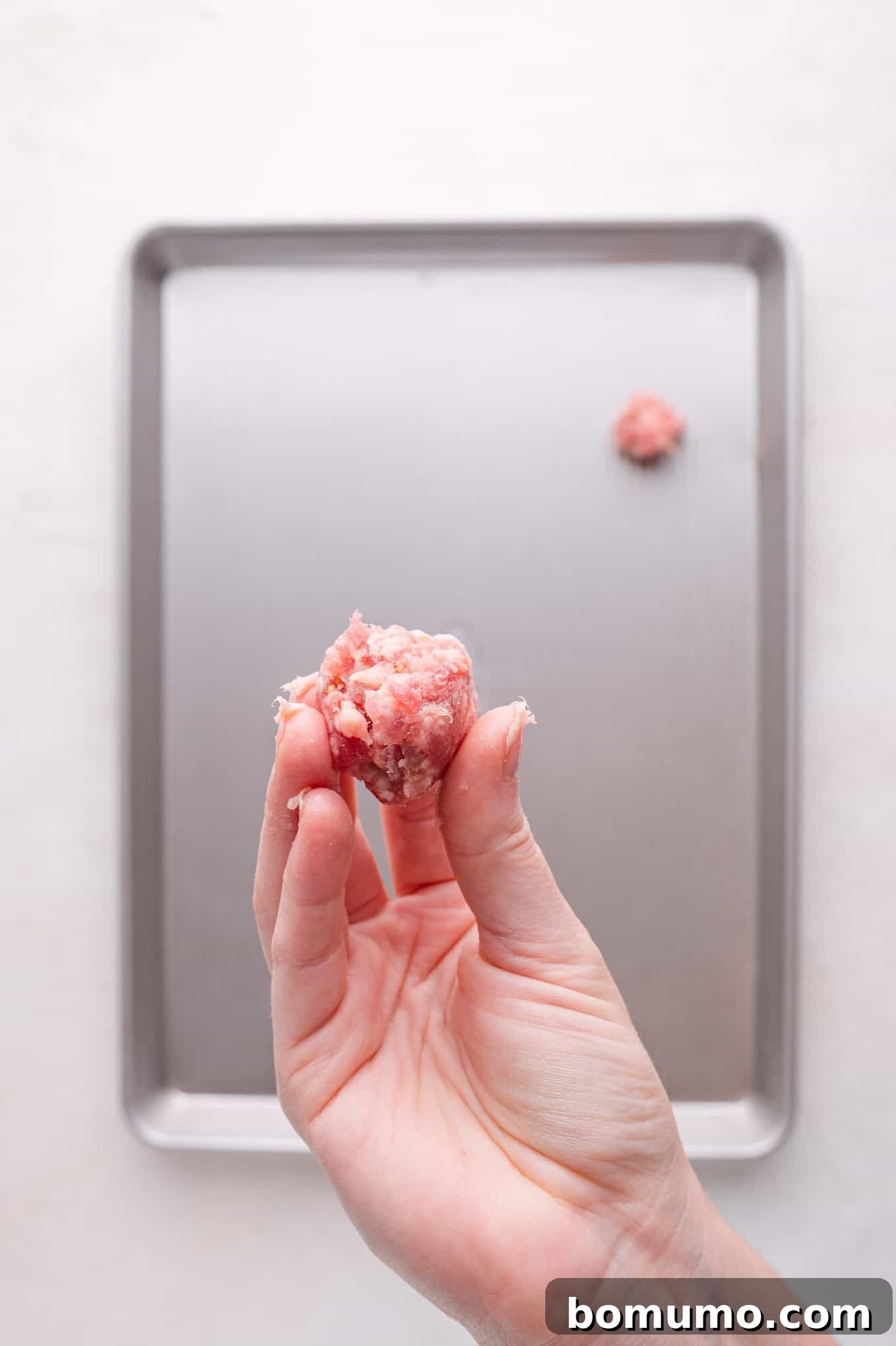A hand gently holds a raw, loosely formed Italian sausage chunk above a metal baking sheet, with several other similar sausage chunks already placed on the tray in the background.
