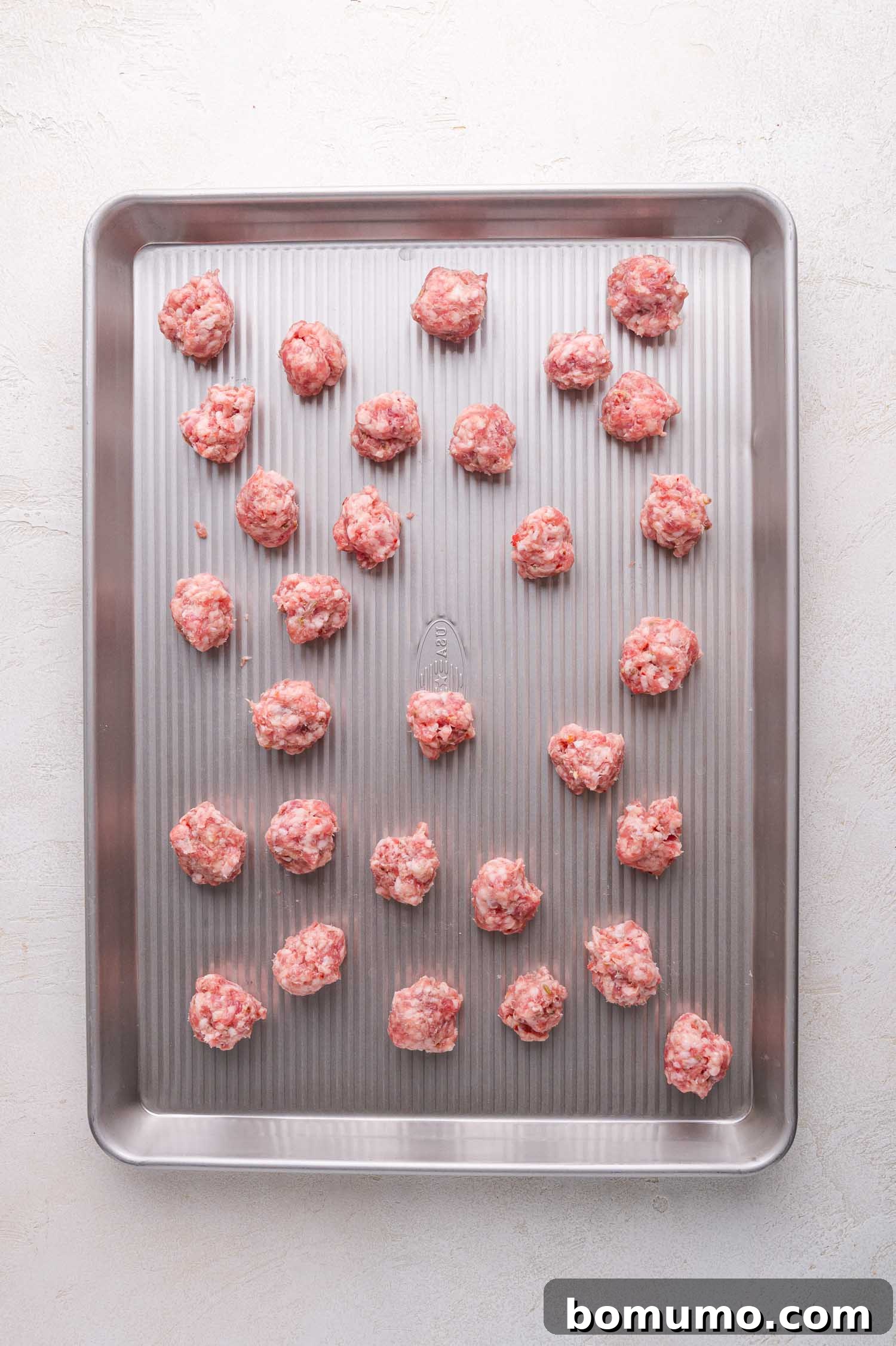 Raw, portioned Italian sausage pieces are spread out on a metal baking sheet, ready for the next step in the cooking process.