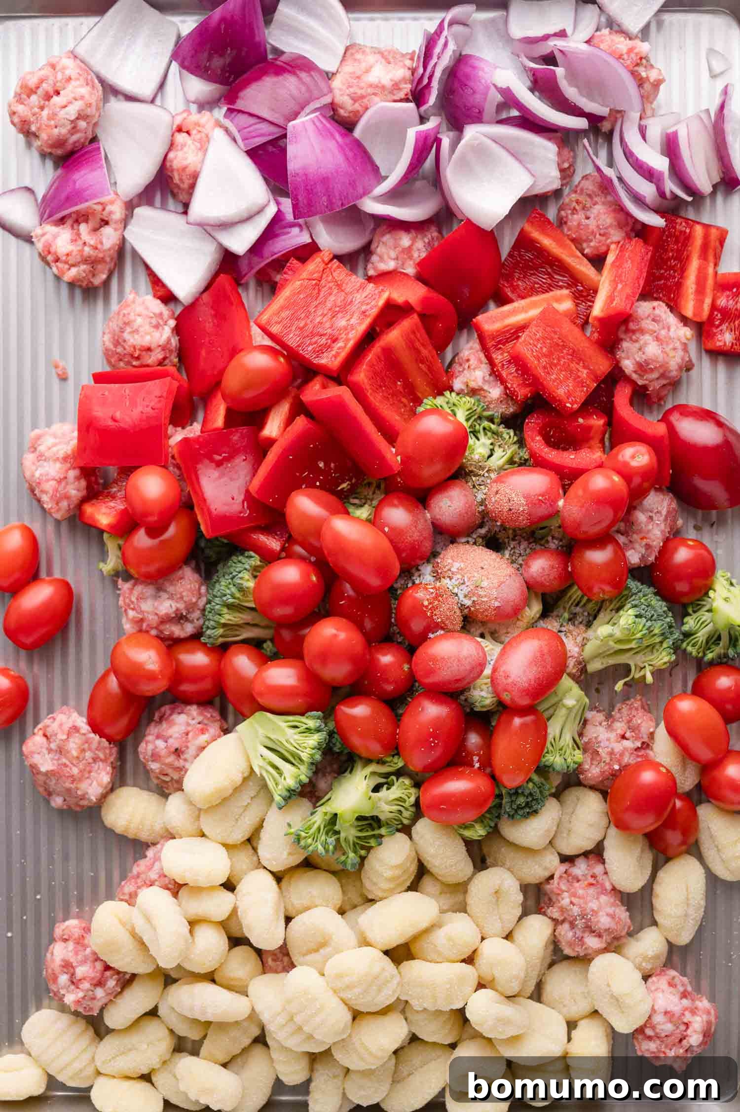 Uncooked gnocchi, vibrant broccoli florets, juicy cherry tomatoes, crisp red bell peppers, and red onion pieces are arranged alongside raw Italian sausage chunks on a spacious baking sheet, coated in olive oil and seasonings.