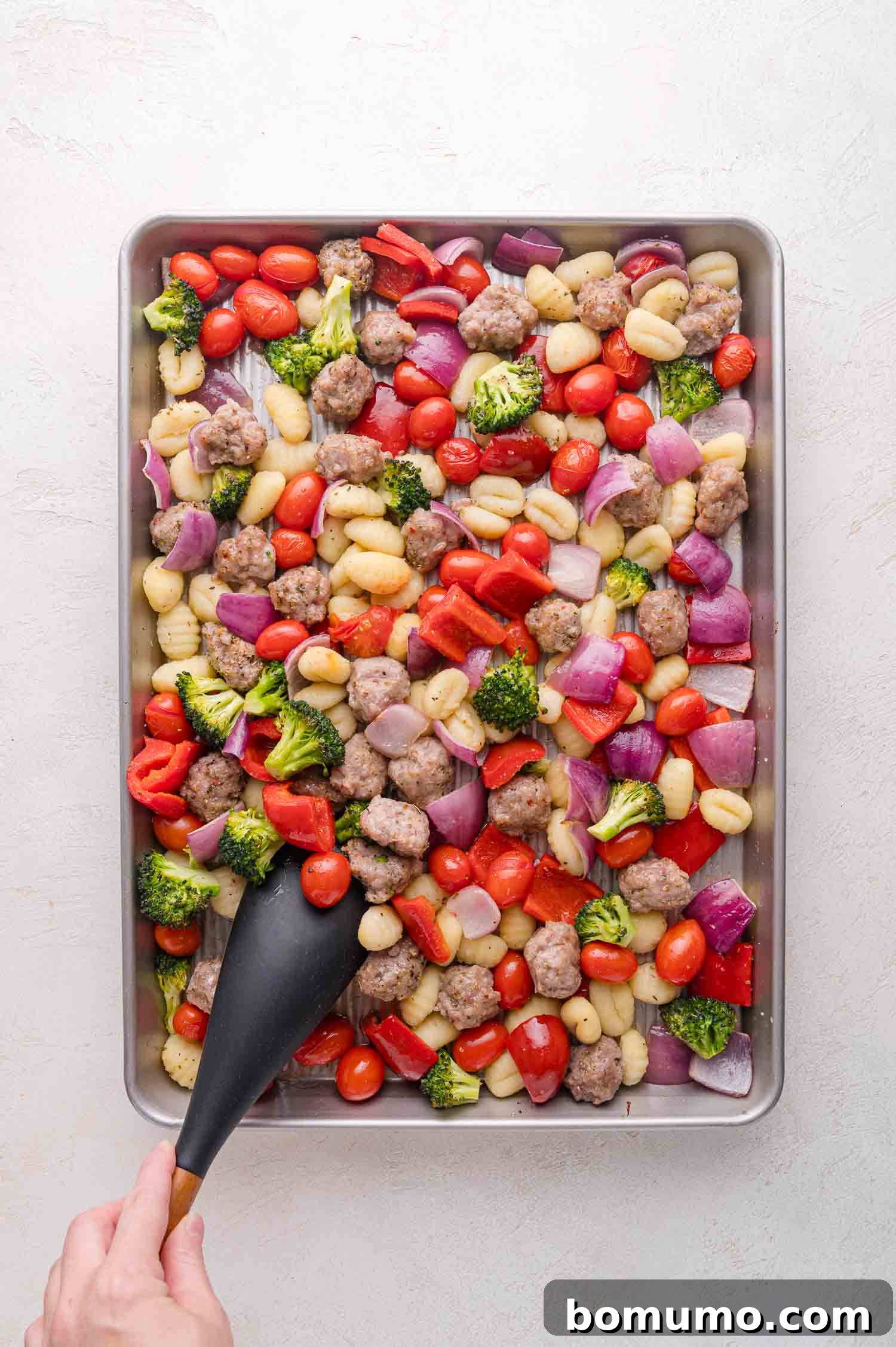 A sheet pan loaded with gnocchi, cherry tomatoes, broccoli, red bell peppers, red onions, and Italian sausage pieces, with a hand holding a black spatula, ready to stir the ingredients during roasting.