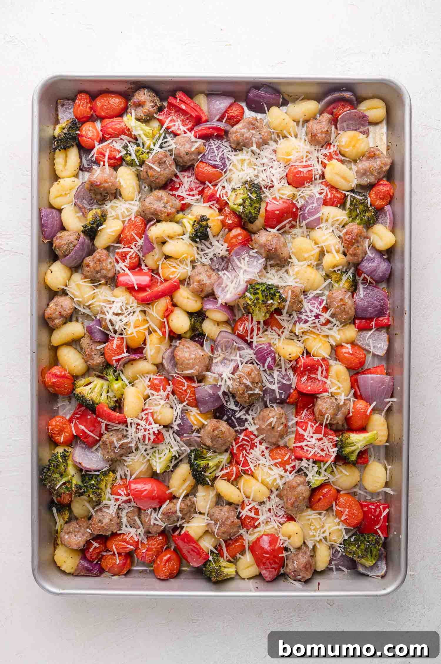 A sheet pan filled with golden-brown gnocchi, cooked Italian sausage, blistered cherry tomatoes, tender red onion, crisp broccoli, and sweet red bell peppers, topped with a generous sprinkle of grated Parmesan cheese, ready to be melted.