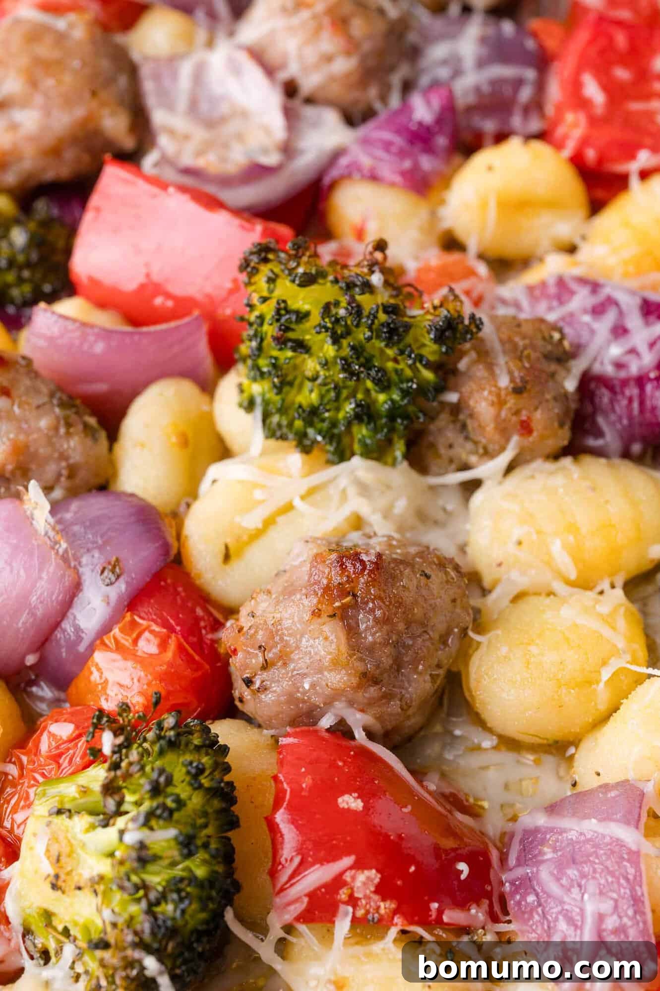 Close-up view of the finished sheet pan gnocchi dish, showcasing crispy gnocchi, savory sausage, perfectly roasted broccoli, red onion, cherry tomatoes, and red bell pepper, all blanketed under a layer of beautifully melted shredded Parmesan cheese.