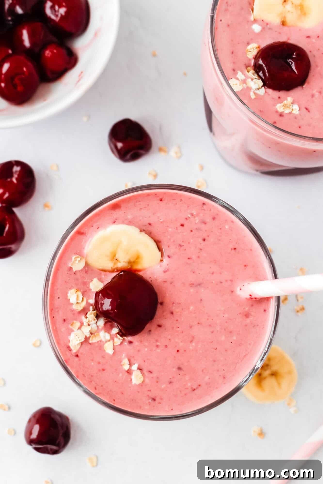 A vibrant pink cherry smoothie in a tall glass, elegantly garnished with a fresh cherry, delicate banana slices, and a sprinkle of wholesome oats. A striped straw invites a sip, while scattered whole cherries and loose oats adorn the table nearby.