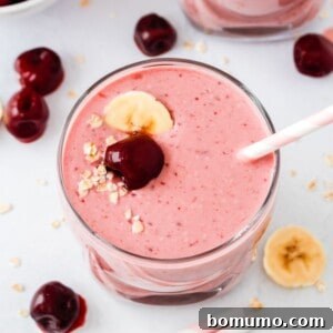 A creamy pink cherry smoothie in a glass, topped with banana slices, a fresh cherry, and oats. A striped straw is present, with scattered cherries and banana slices on the side.