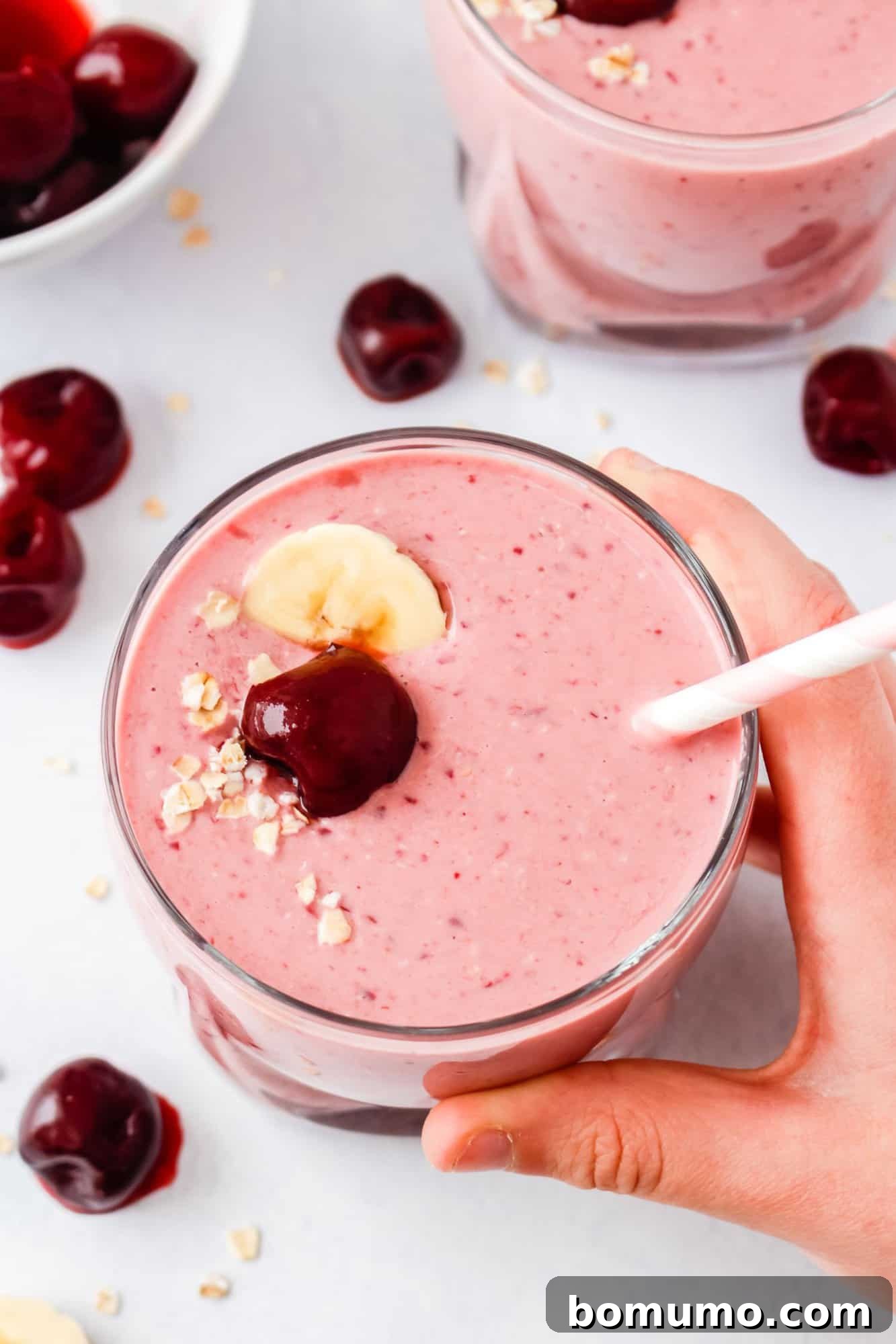 A hand gently holds a tall glass filled with a bright pink cherry smoothie, artfully topped with a fresh cherry, a perfect banana slice, and a scattering of oats. A striped straw protrudes invitingly, while loose cherries are casually strewn on the clean white surface below.