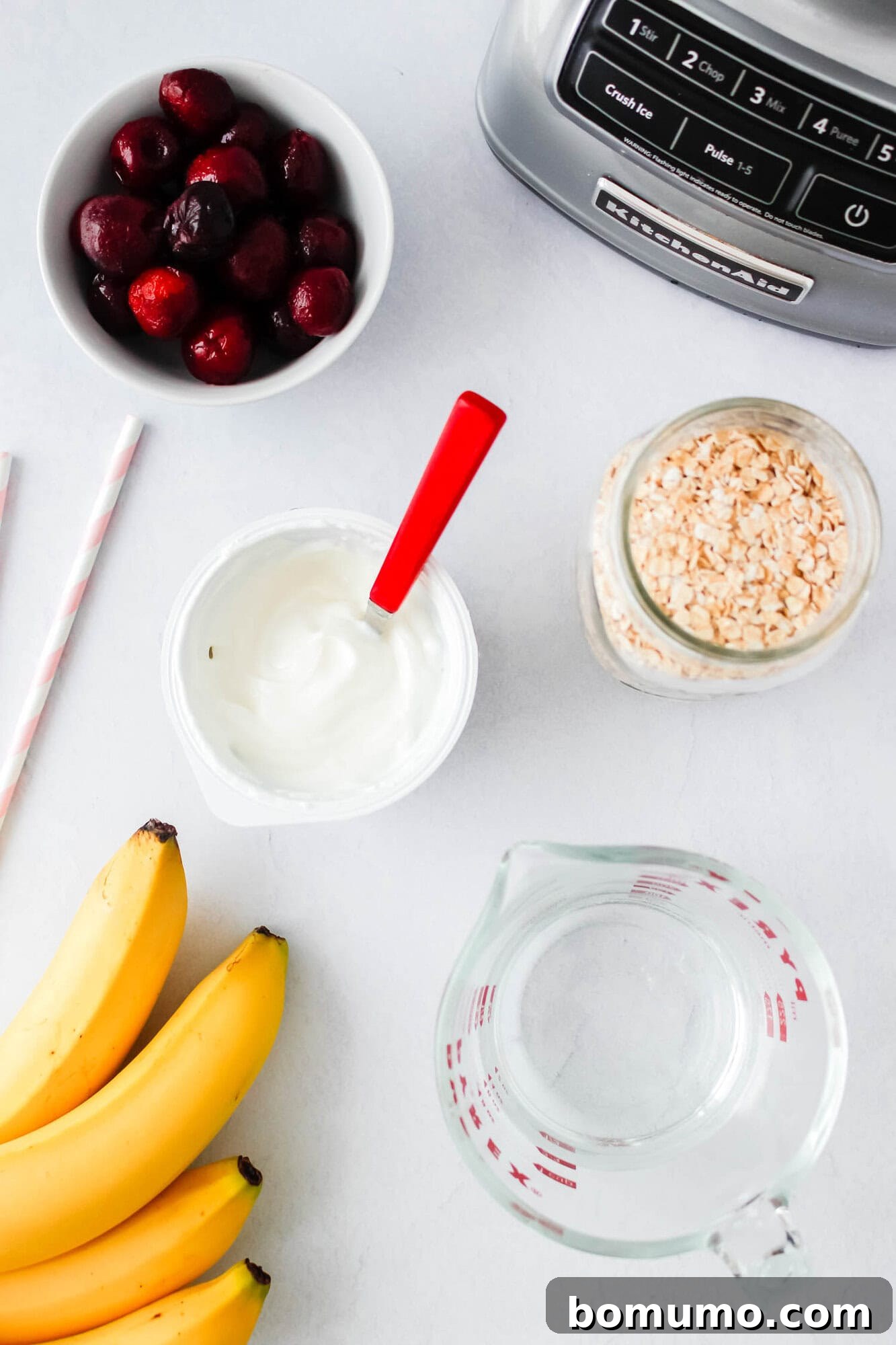 A selection of fresh and frozen smoothie ingredients artfully arranged on a kitchen countertop: a bowl of ripe cherries, banana slices, a container of creamy vanilla Greek yogurt with a vibrant red spoon, a glass measuring cup for liquid, and a jar of rolled oats. A blender is partially visible in the background, signaling the start of the preparation.