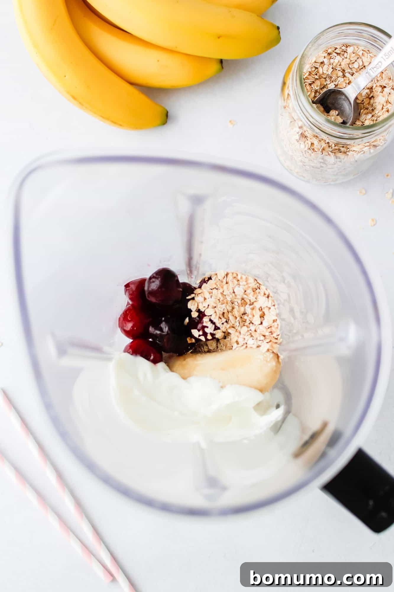 The initial stage of smoothie preparation: a blender jug contains a vibrant mix of creamy Greek yogurt, bright red cherries, yellow banana slices, and a scoop of rolled oats. Beside the blender, fresh bananas and a jar of oats with a spoon are arranged on a clean white surface, illustrating the raw ingredients.