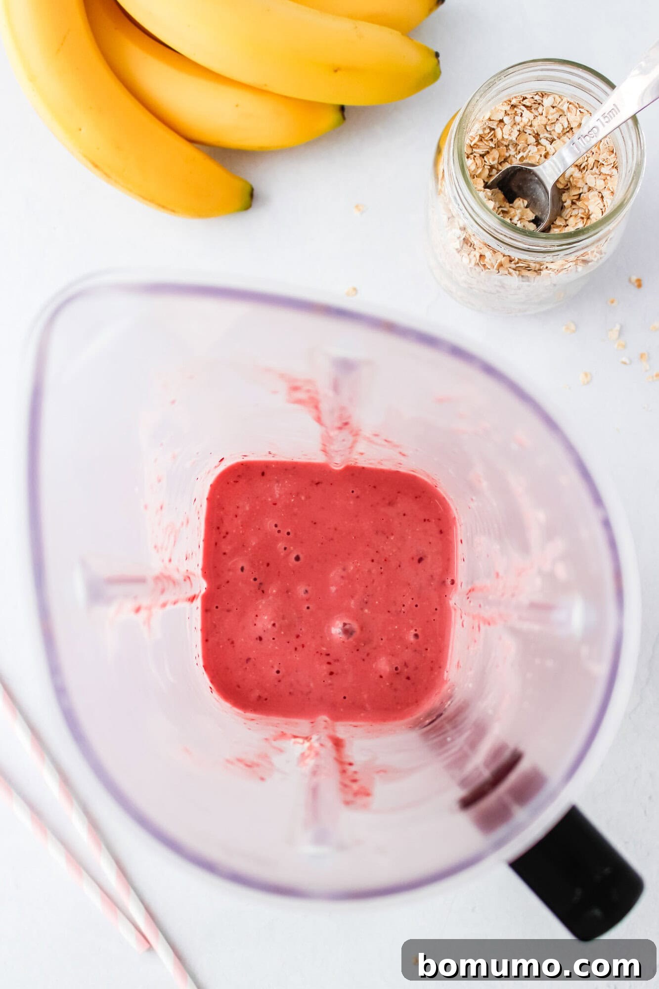 A high-speed blender, now filled with a perfectly smooth and creamy pink cherry smoothie, rests on a pristine white surface. Nearby, a fresh banana, a jar of oats with a spoon, and two striped straws are neatly placed, symbolizing the completion of the blending process and readiness for enjoyment.