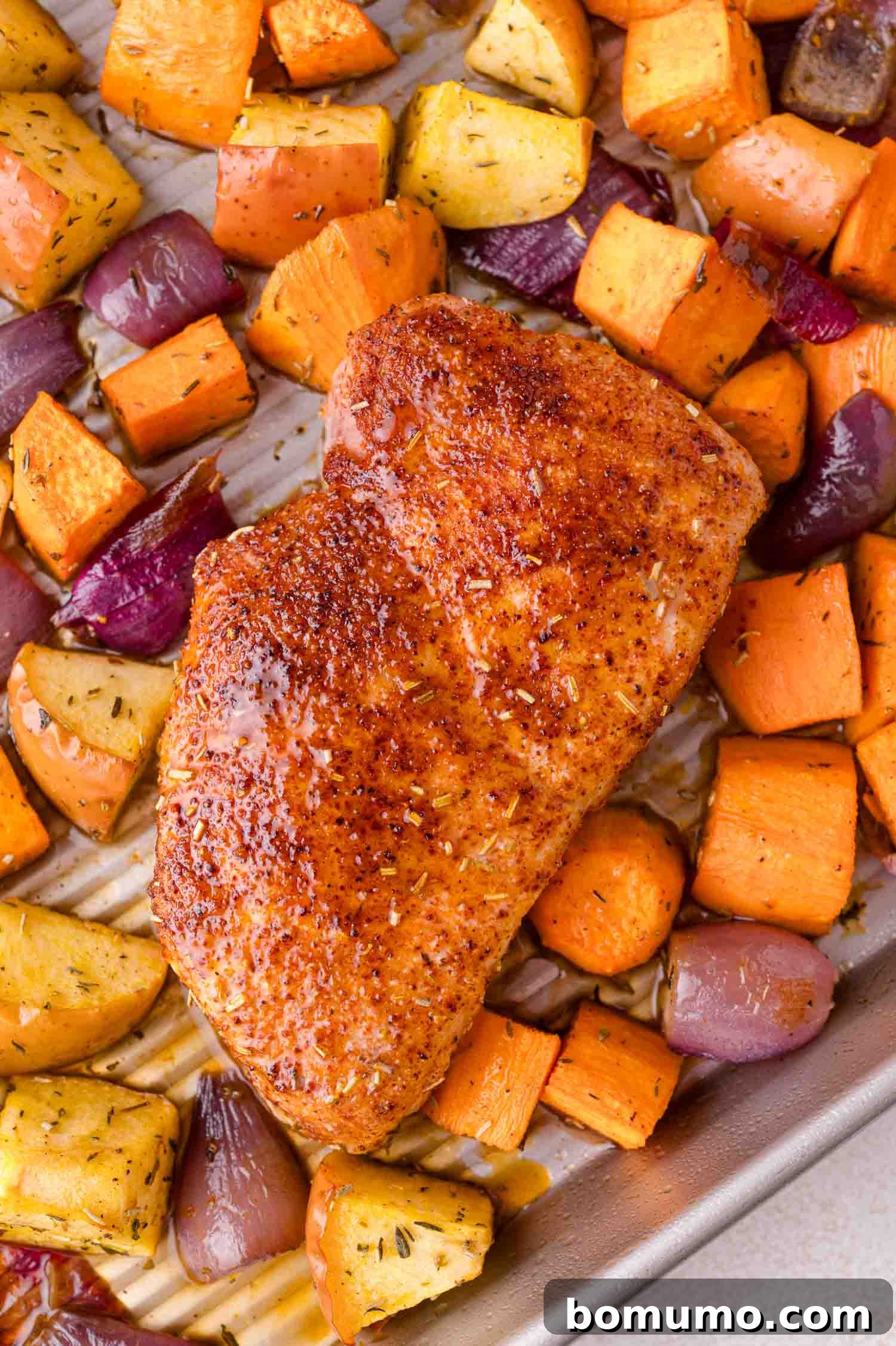 A perfectly seasoned pork chop, roasted to a golden brown, resting on a baking sheet alongside tender sweet potatoes, caramelized apples, and delicate red onion slices.