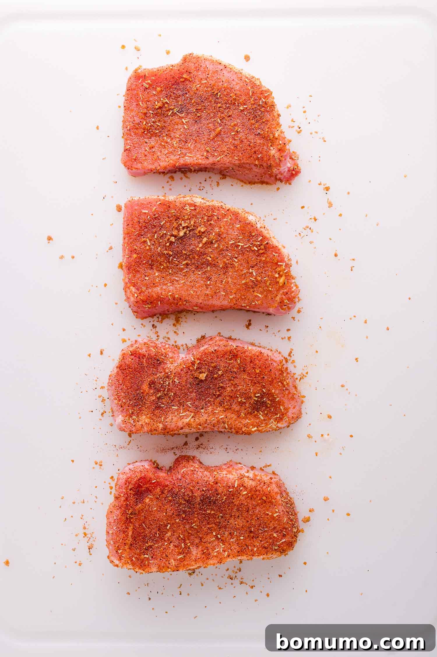 Four raw pork chops, generously coated with a dry seasoning rub, are neatly arranged in a vertical line on a clean white surface, with a scattering of spices around them, indicating they are fully prepared for cooking.