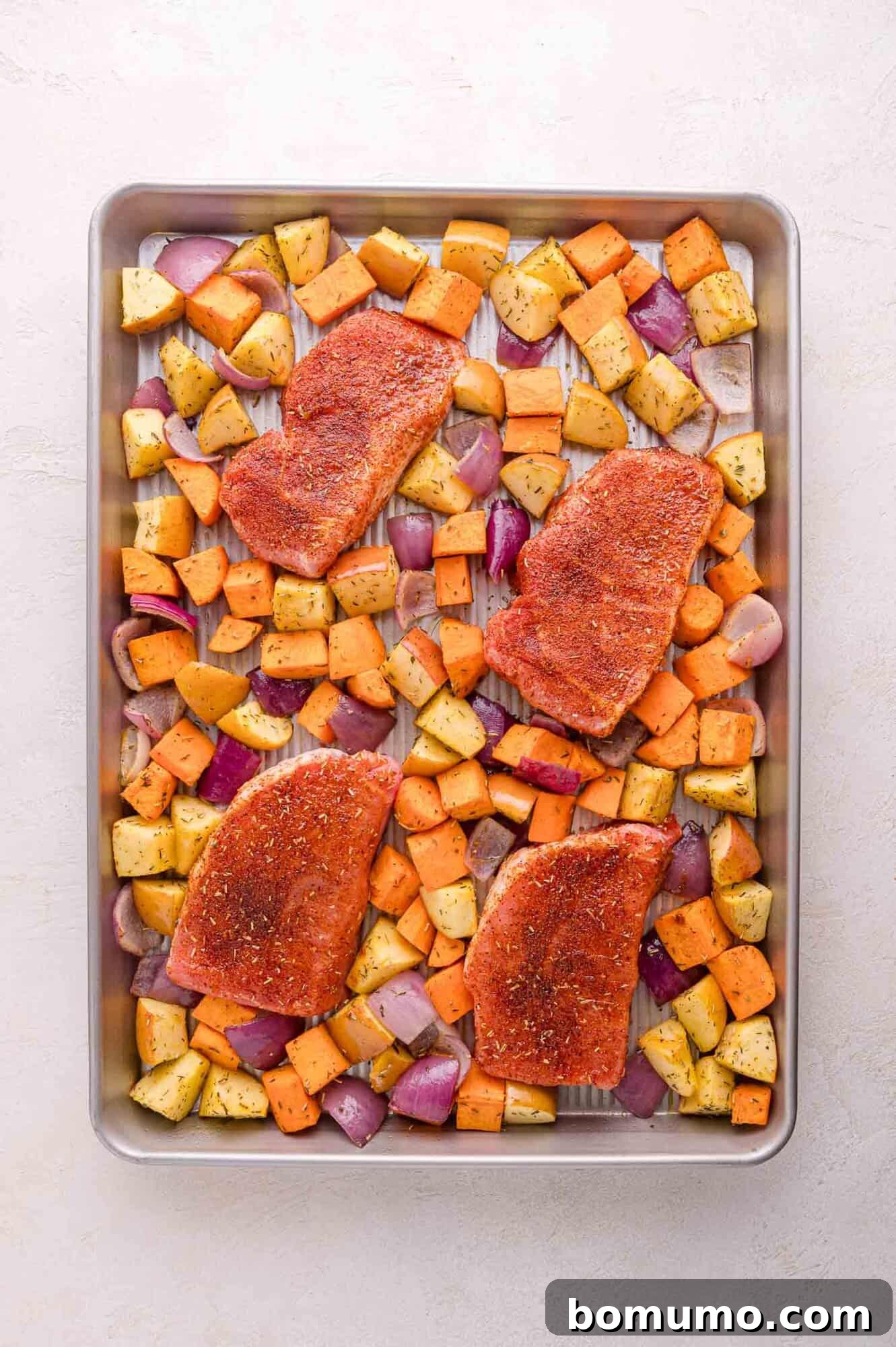 A sheet pan with four seasoned raw pork chops strategically placed among a colorful bed of diced sweet potatoes, crisp apples, and red onions, all ready to be roasted together in the oven for a complete meal.