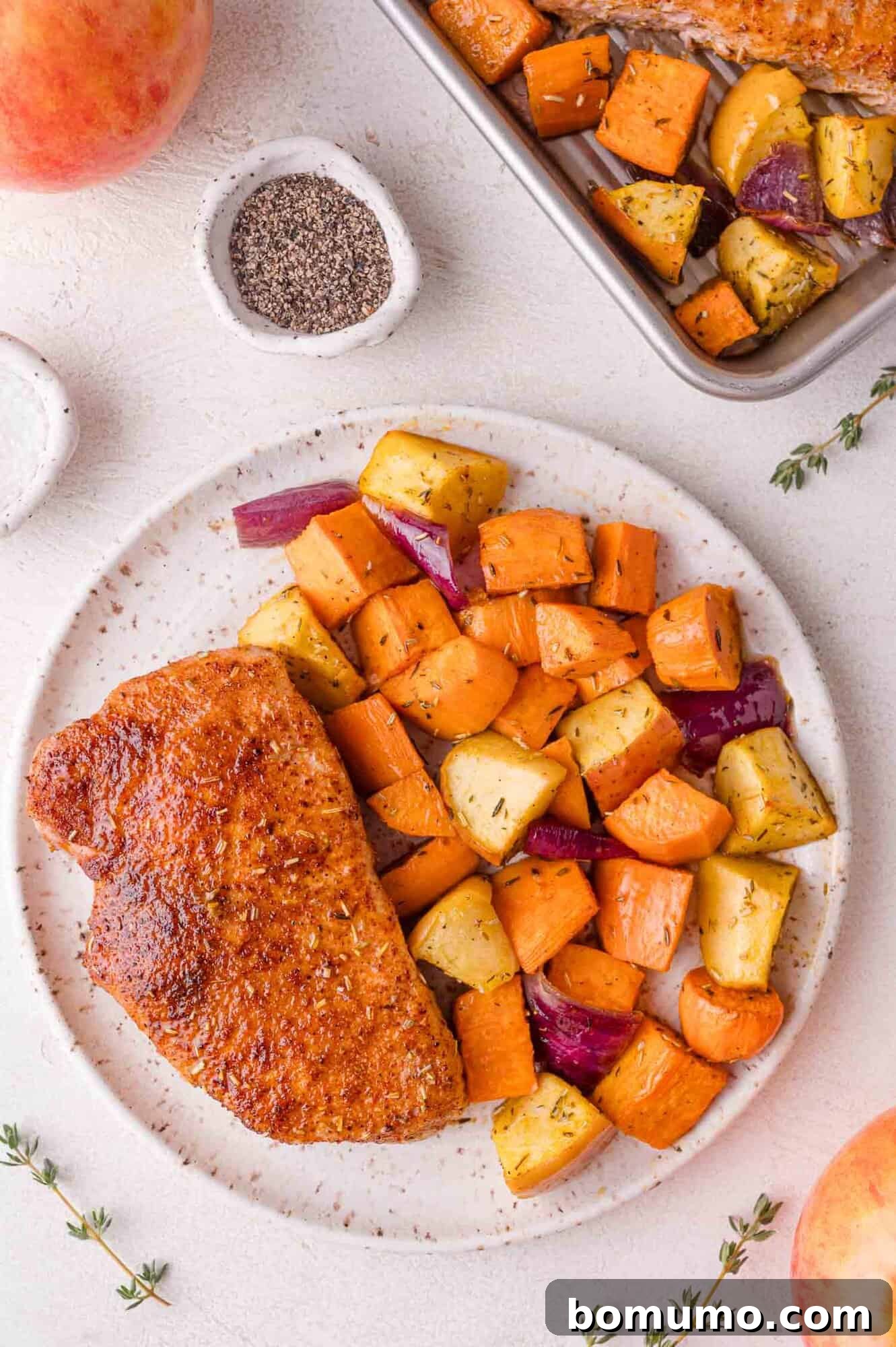 A plate featuring a perfectly seasoned and cooked pork chop, accompanied by a generous portion of roasted vegetables, including tender sweet potatoes, sweet apples, and savory red onions, all resting on a pristine white surface. In the background, a pan holds additional roasted vegetables, suggesting a bountiful meal.
