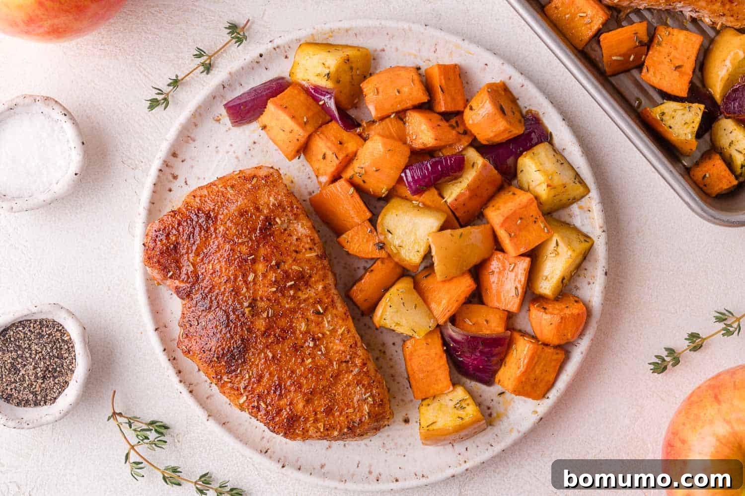 A beautifully plated, seasoned cooked pork chop accompanied by roasted sweet potatoes, red onions, and sweet apples. Garnish of fresh thyme and a sprinkle of salt and pepper elevate the presentation.