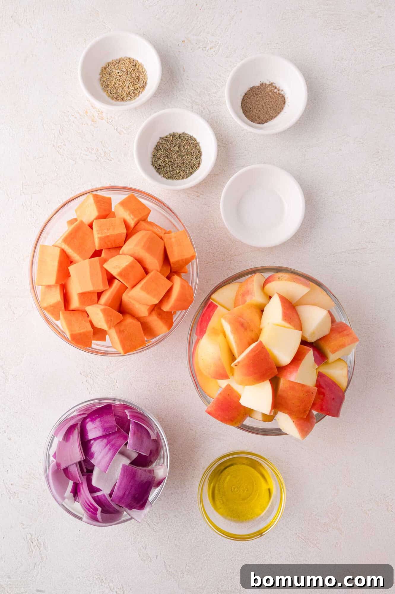Bowls of neatly diced sweet potatoes, freshly chopped apples, and sliced red onion, accompanied by small dishes of olive oil, dried herbs, and various seasonings, all arranged on a light surface, ready for preparation.