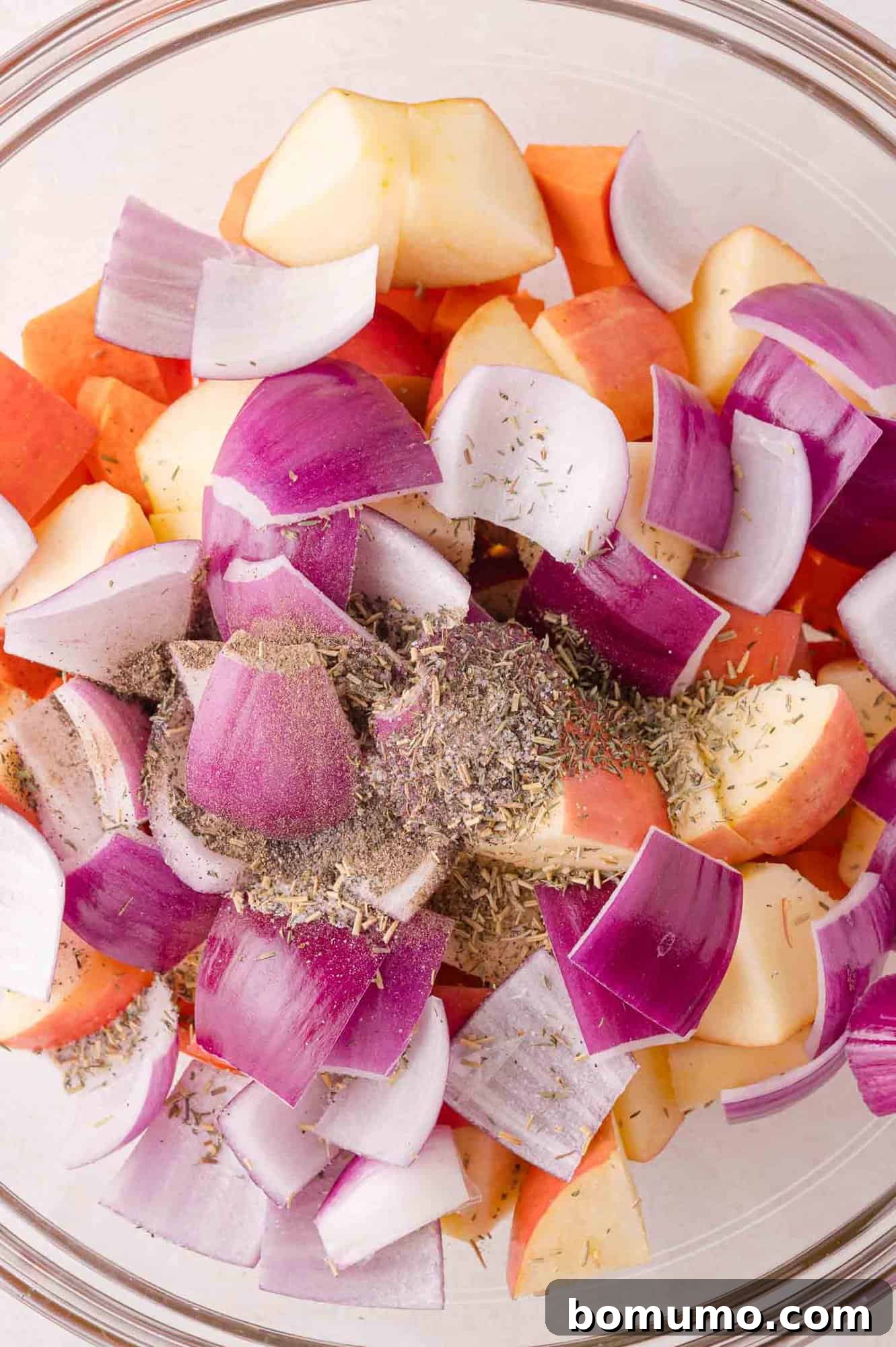 A glass bowl holds a vibrant mix of chopped red onions, crisp apples, and sweet potatoes, visibly seasoned with aromatic dried herbs and a generous grinding of black pepper, awaiting a good toss before roasting.