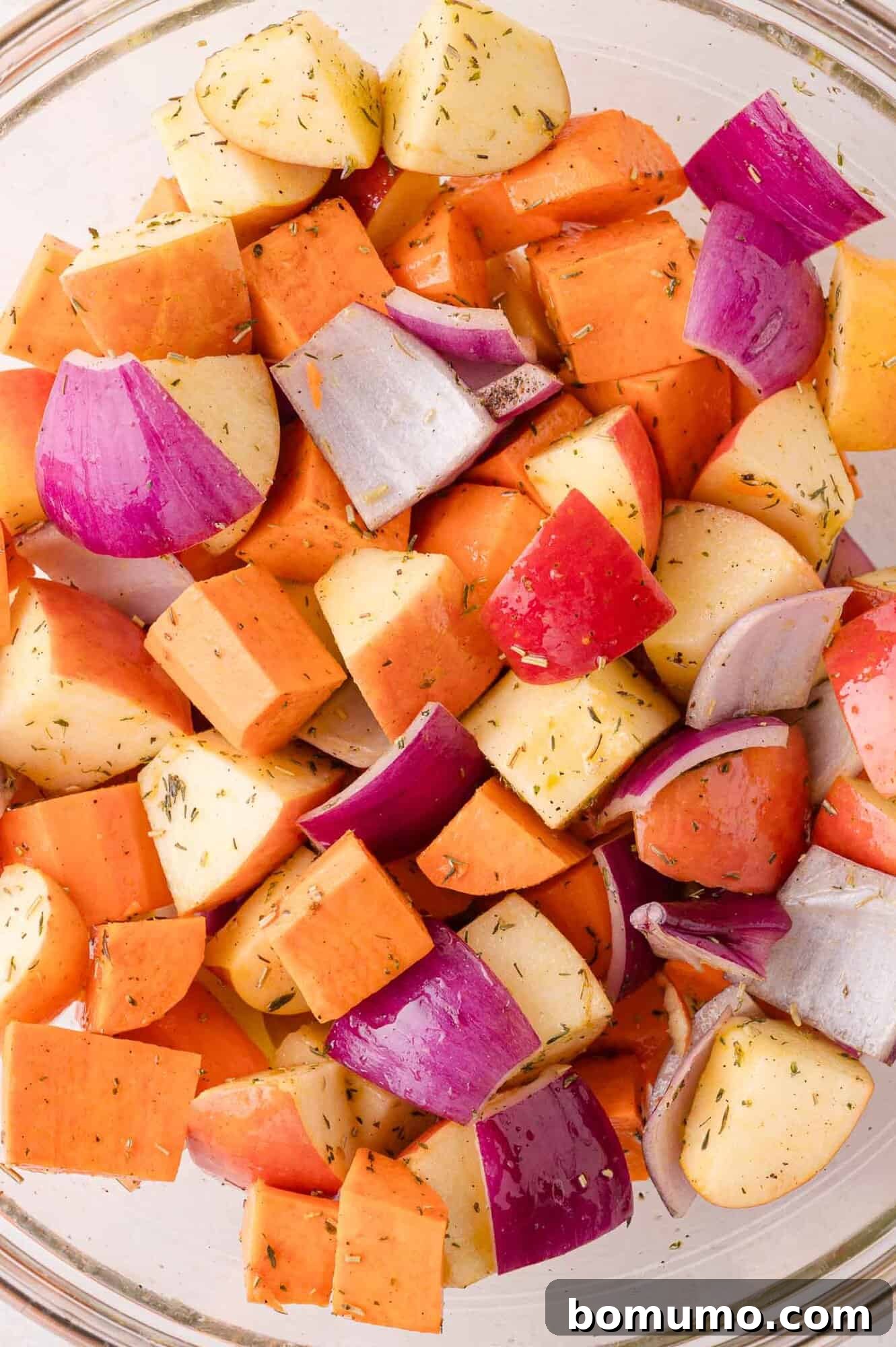 A close-up view of a glass bowl filled with a colorful mixture of chopped sweet potatoes, apples, and red onions, thoroughly coated with fragrant herbs and spices, showcasing their even distribution.