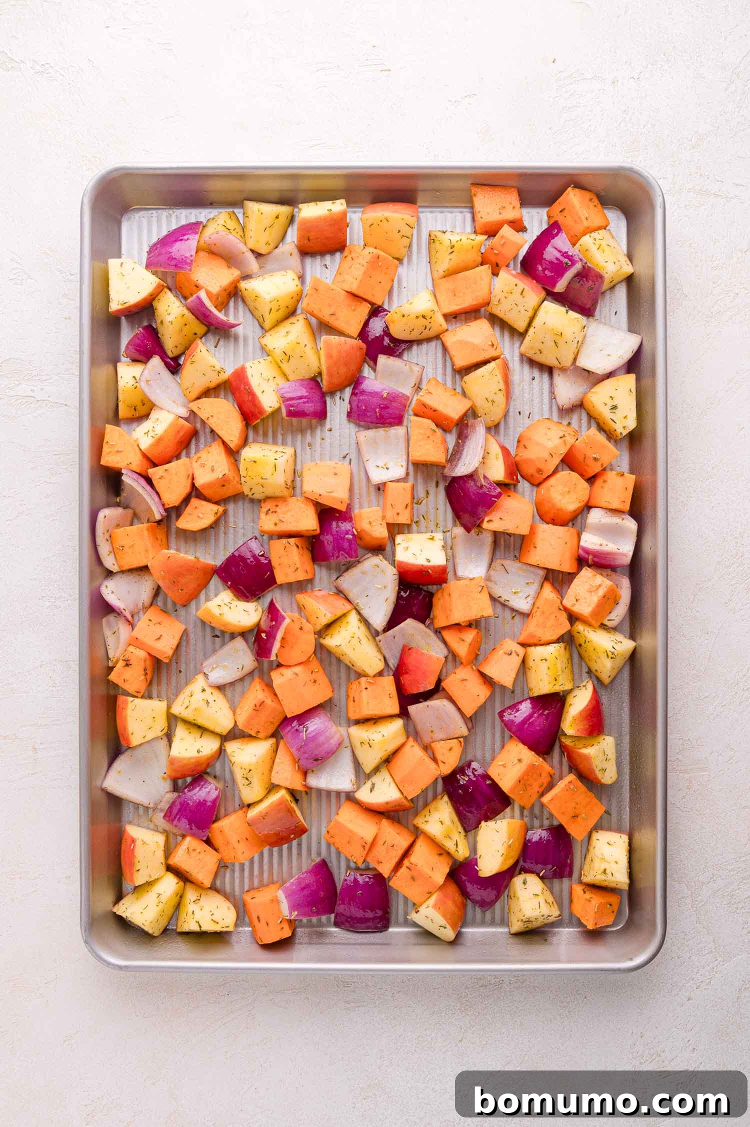 A sheet pan beautifully arranged with a medley of chopped sweet potatoes, red onions, and apples, all evenly seasoned with a blend of dried herbs, poised and ready for the oven to begin roasting.