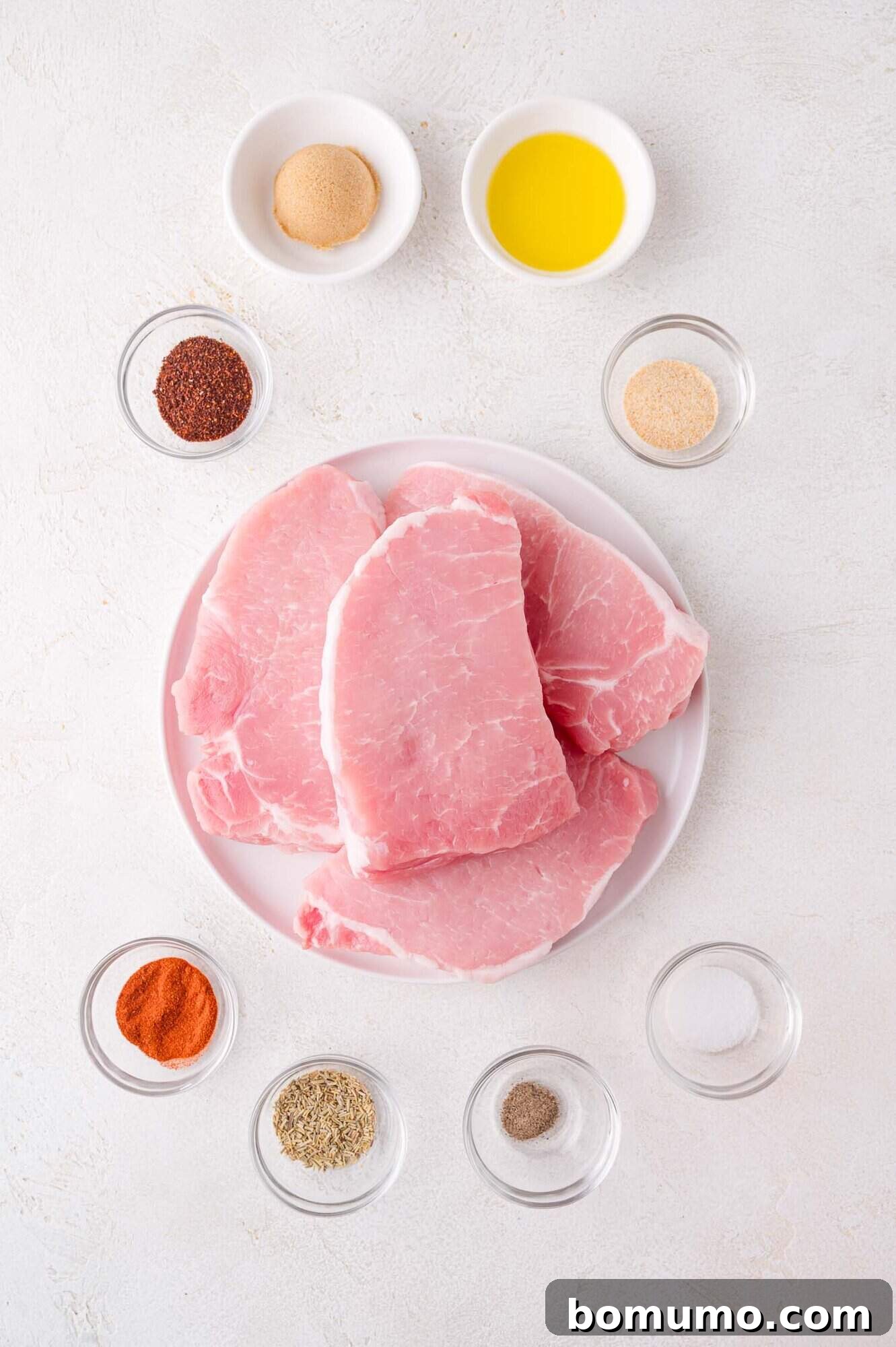 A plate featuring raw pork chops, artfully surrounded by an assortment of small bowls containing various spices, seasonings, olive oil, and brown sugar, all set on a pristine white surface, ready for the seasoning process.