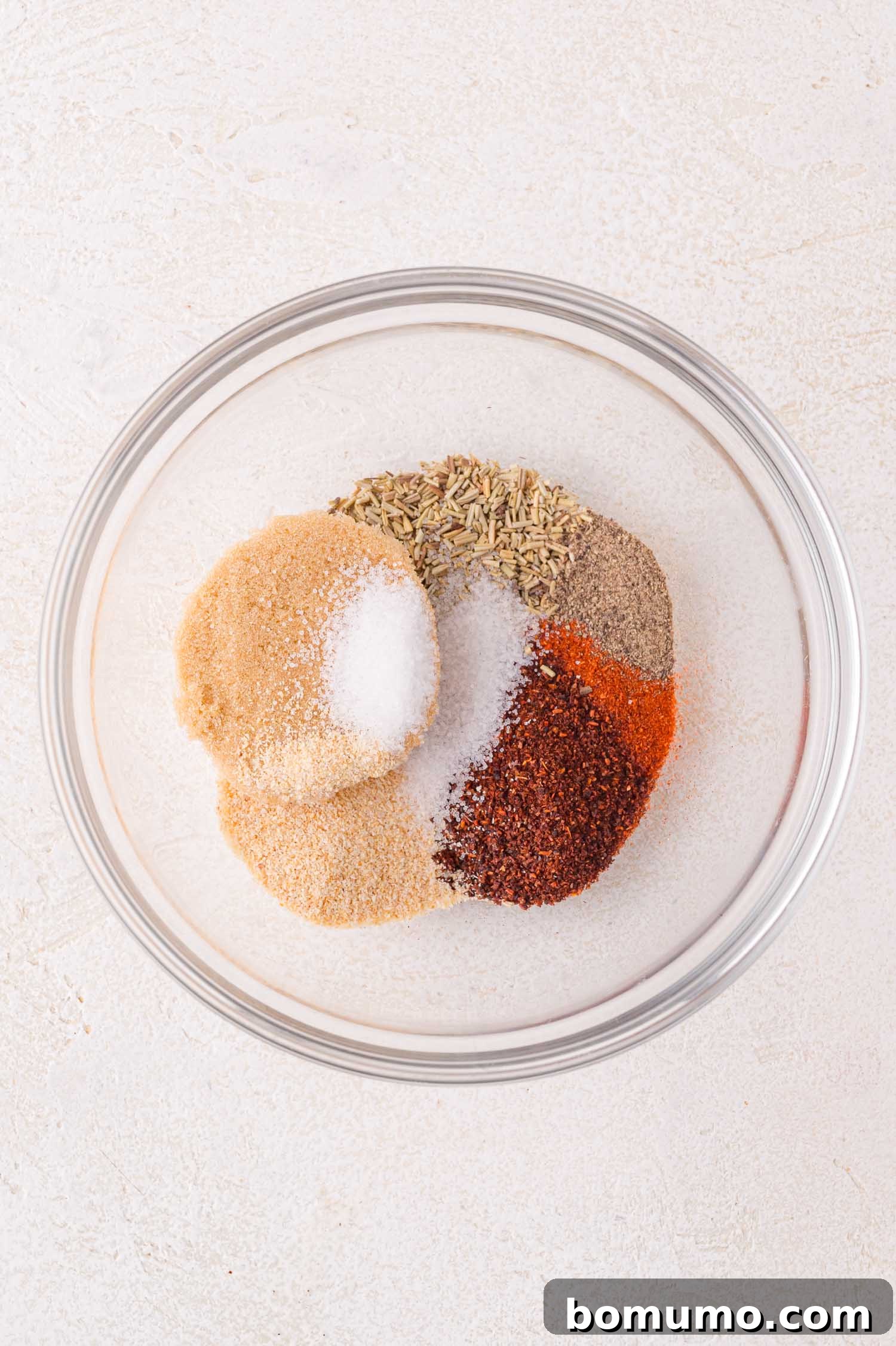 A clear glass bowl showcasing distinct piles of various ground spices and seasonings, including salt, black pepper, vibrant paprika, earthy cumin, and potent garlic powder, all arranged on a light-colored surface, highlighting the individual components of the rub.