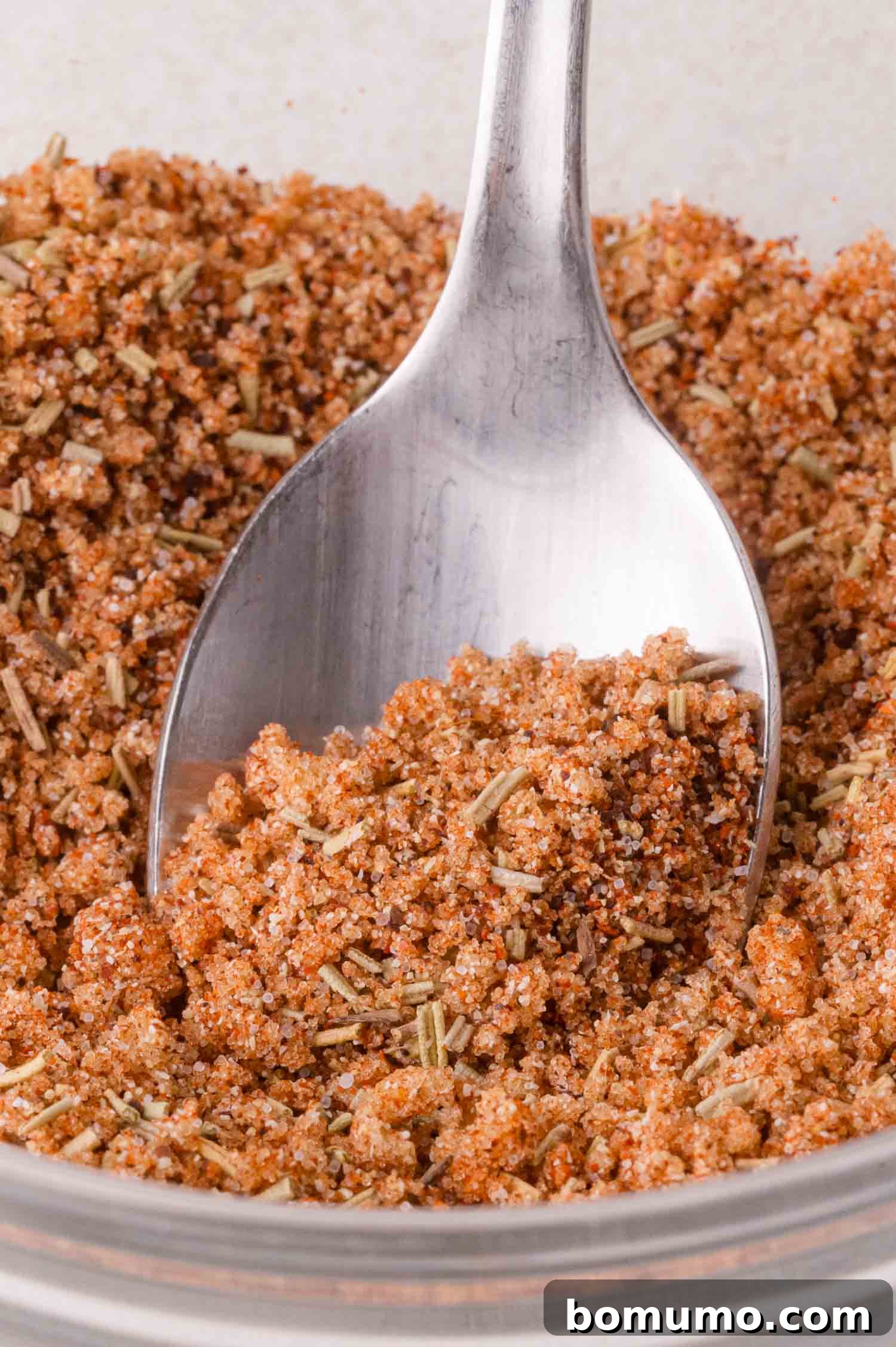 A close-up shot capturing a spoon in the act of scooping a fragrant mixture of ground spices and dried herbs from a glass container, illustrating the preparation of the seasoning blend.